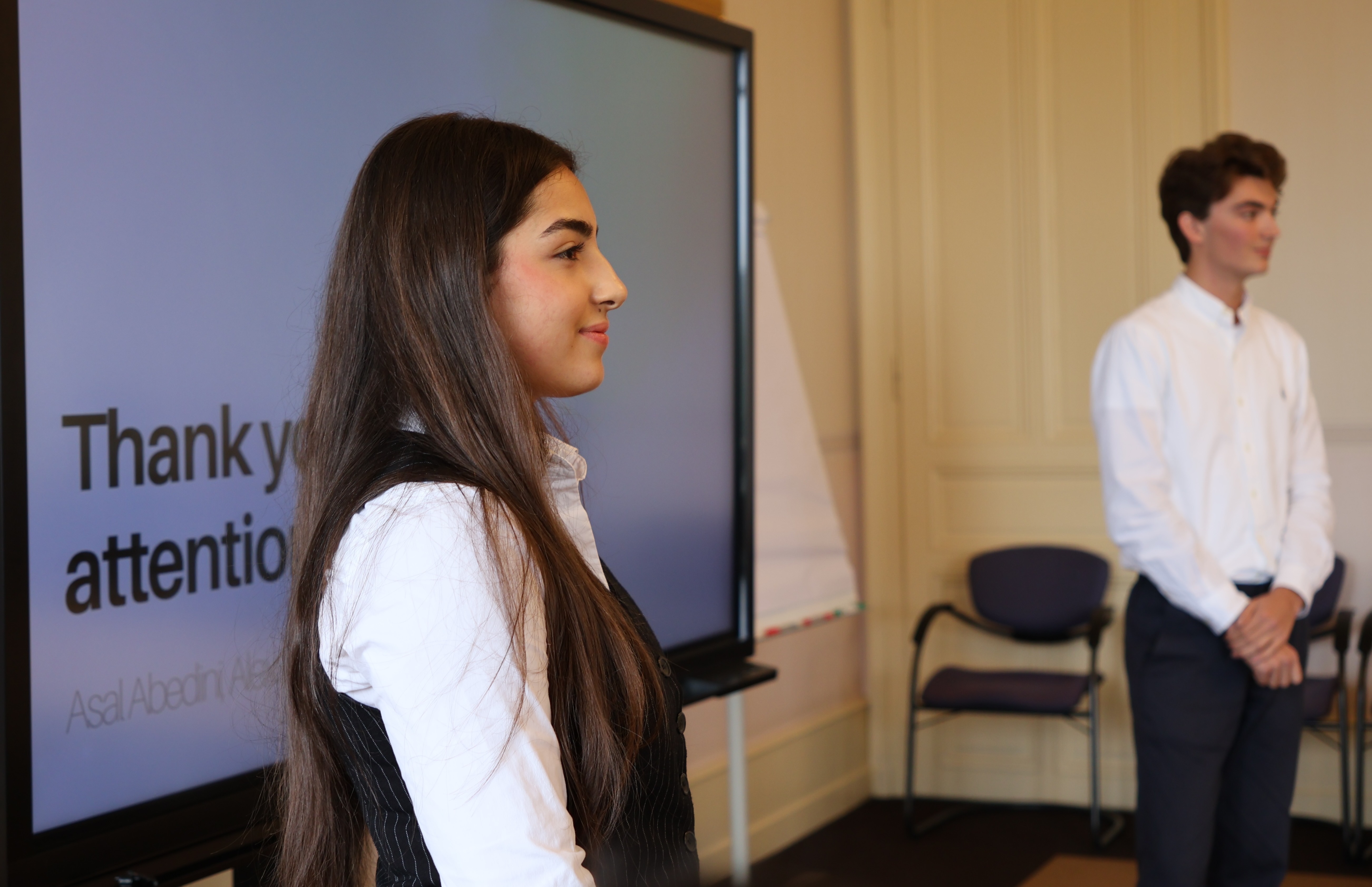 Young woman in white shirt and black vest standing in front of a screen showing 'Thank you for your attention.'