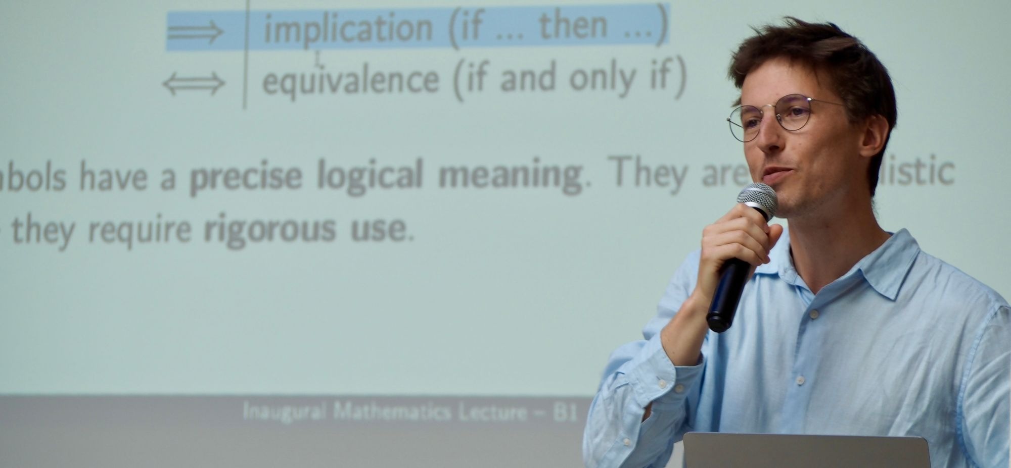 Man with glasses speaking into a microphone in front of a projected slide about logical implication and equivalence.