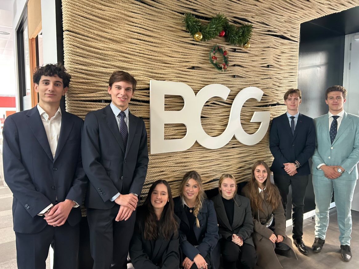 Seven young professionals dressed in business attire posing in front of a wall with a large BCG logo and holiday decorations.