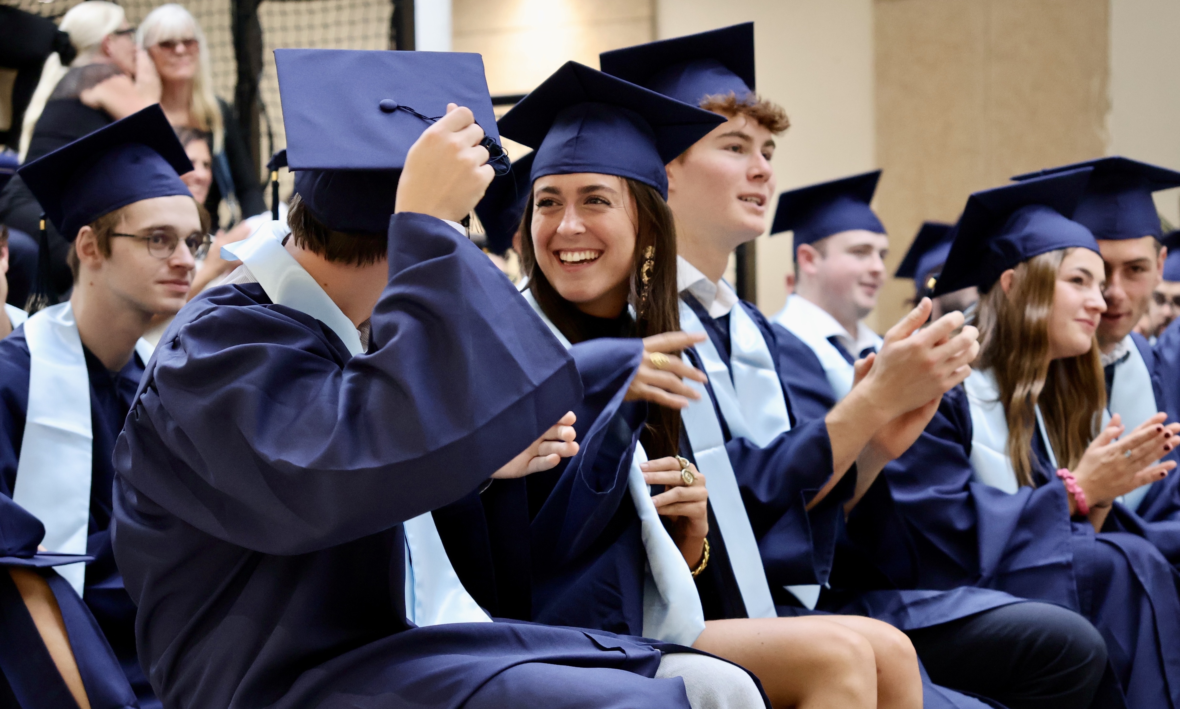 Group of smiling graduates in blue caps and gowns sitting and interacting during a ceremony.