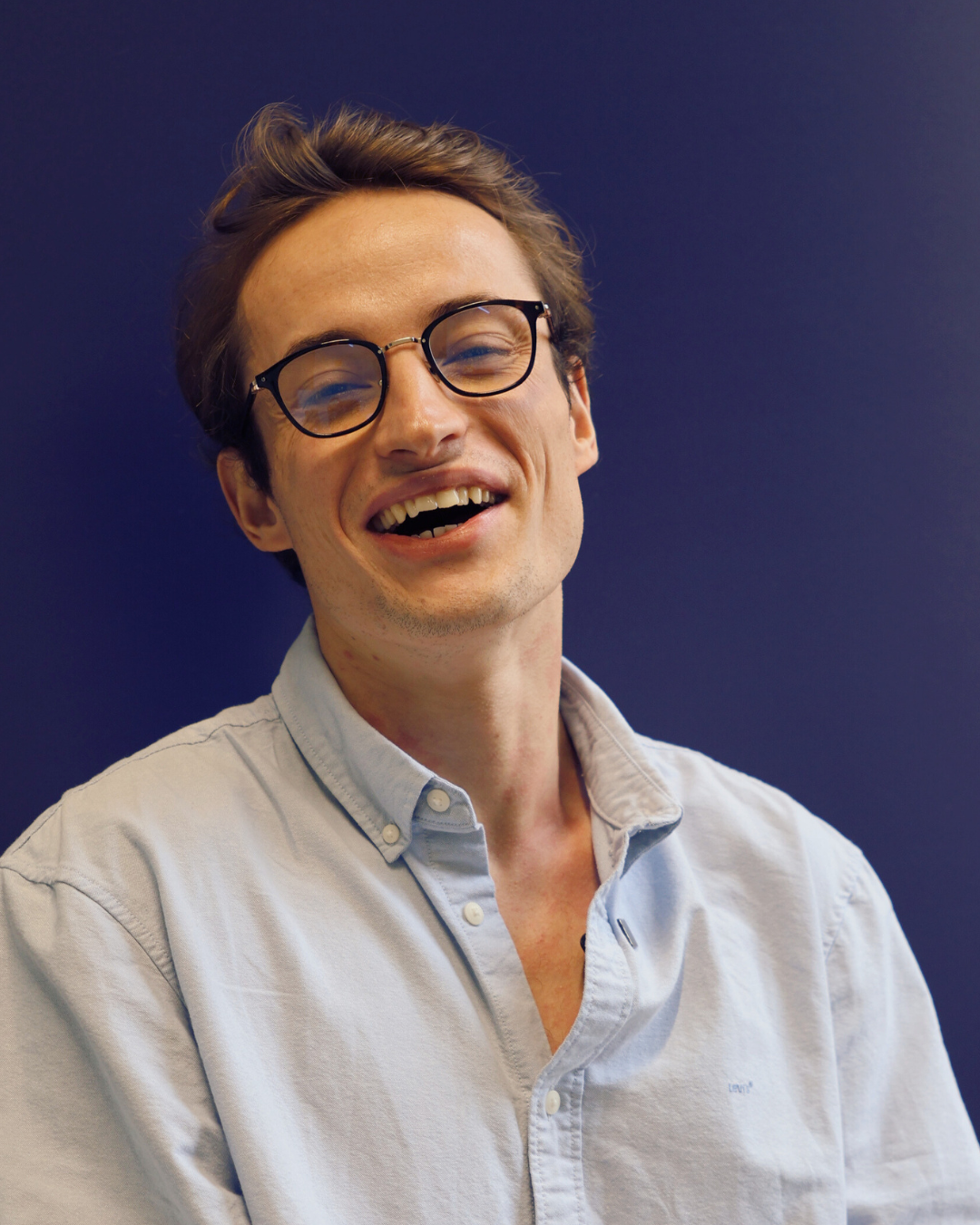 Young man with glasses smiling and laughing against a dark blue background.