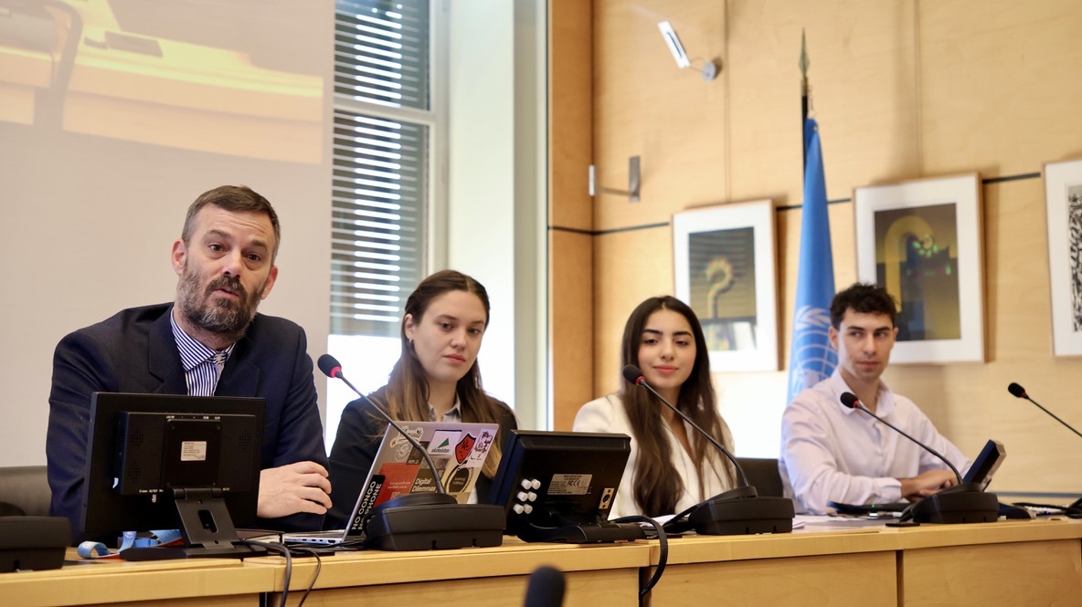 Four people seated at a conference table with microphones and laptops in front of them during a formal meeting.