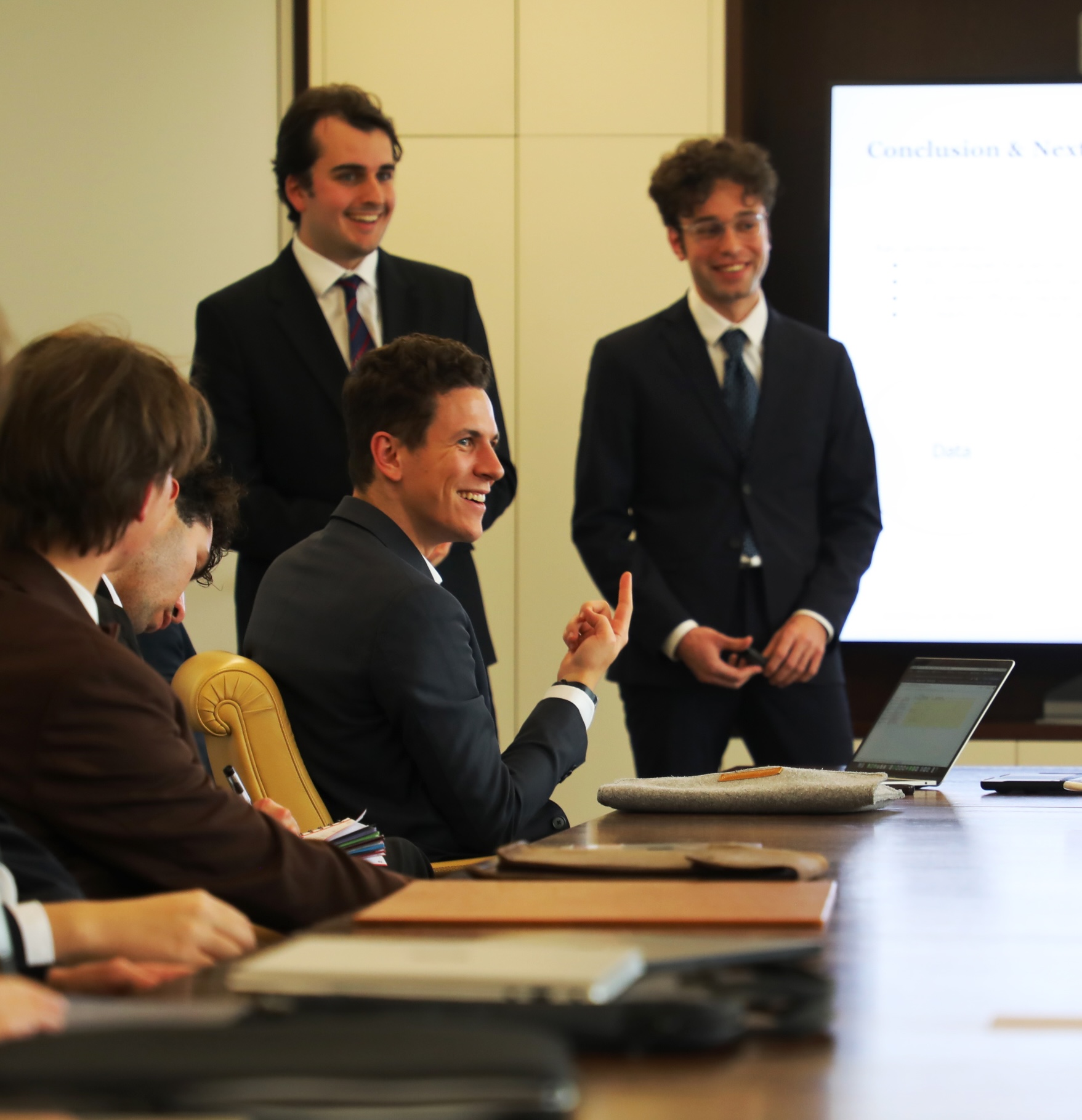 Young professionals in suits engaged in a business meeting with a laptop and presentation screen in the background.