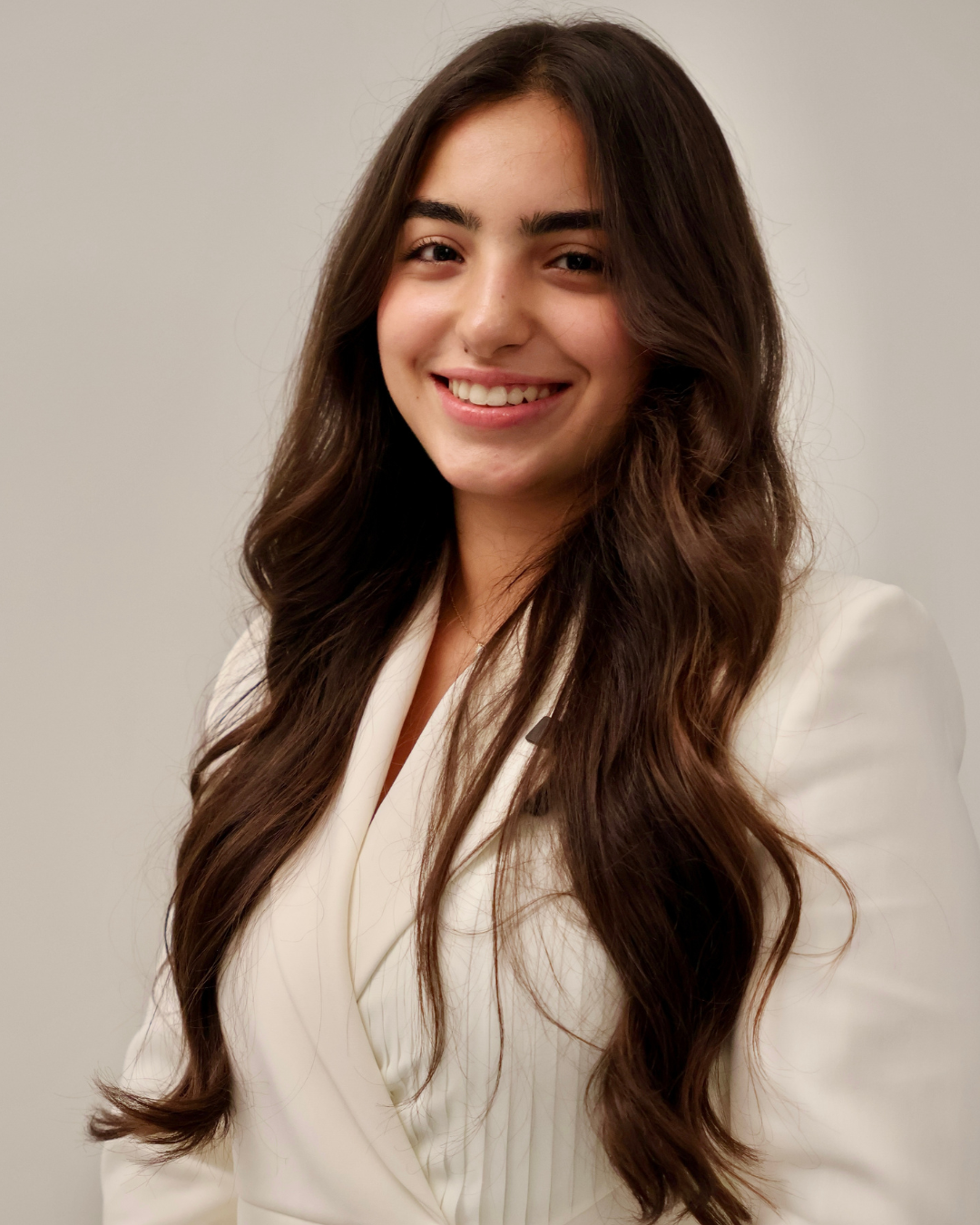 Young woman with long wavy brown hair wearing a white blazer, smiling against a plain light background.