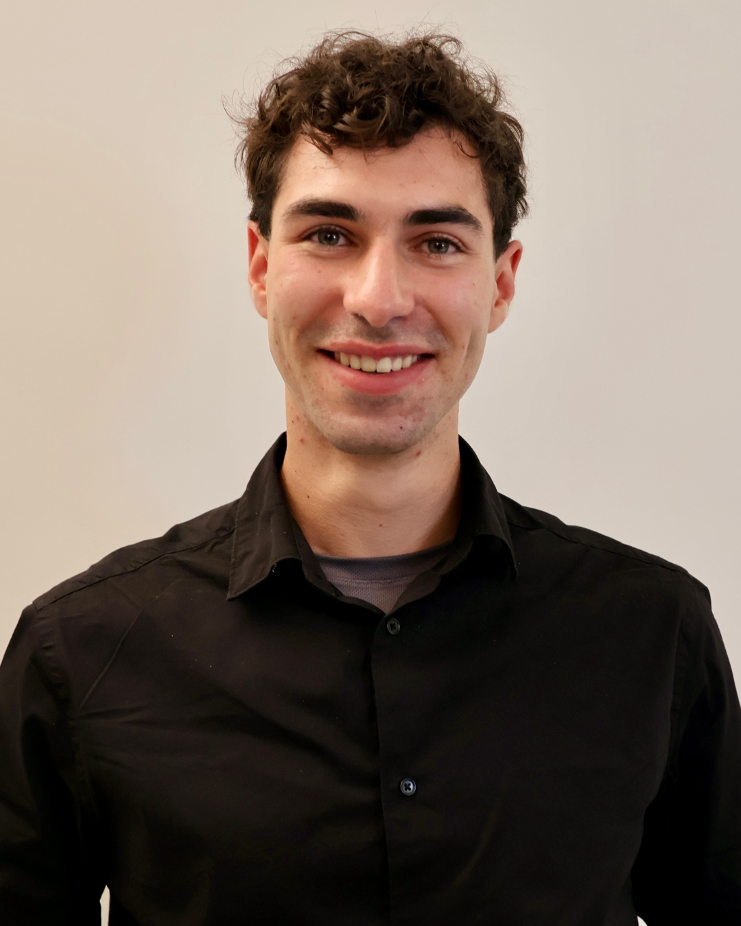 Smiling man with curly dark hair wearing a black collared shirt against a plain light background.