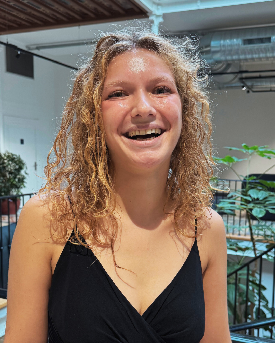 Smiling young woman with curly blonde hair wearing a black sleeveless top in an indoor setting with plants.