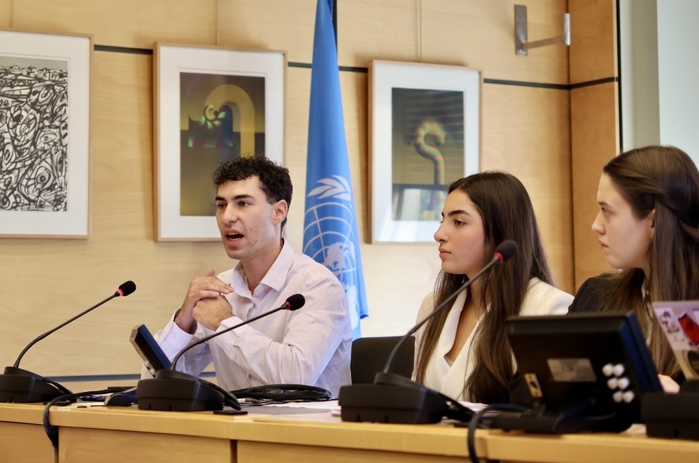 Three young adults sitting at a conference table with microphones, one man speaking, with a United Nations flag and framed abstract artwork in the background.