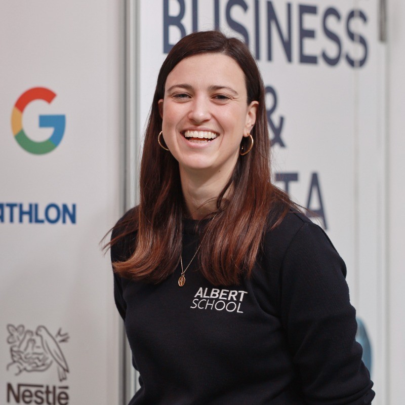 Smiling woman with long brown hair wearing a black Albert School sweatshirt and gold hoop earrings.