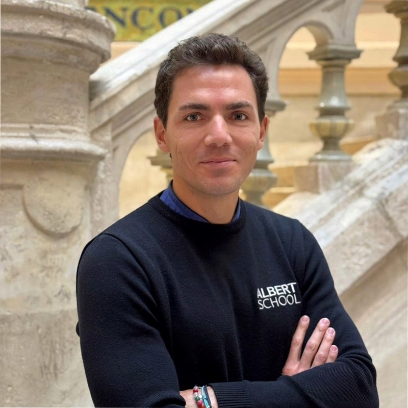 Man with curly dark hair wearing a black sweater with Albert School logo, standing with arms crossed in front of stone staircase.