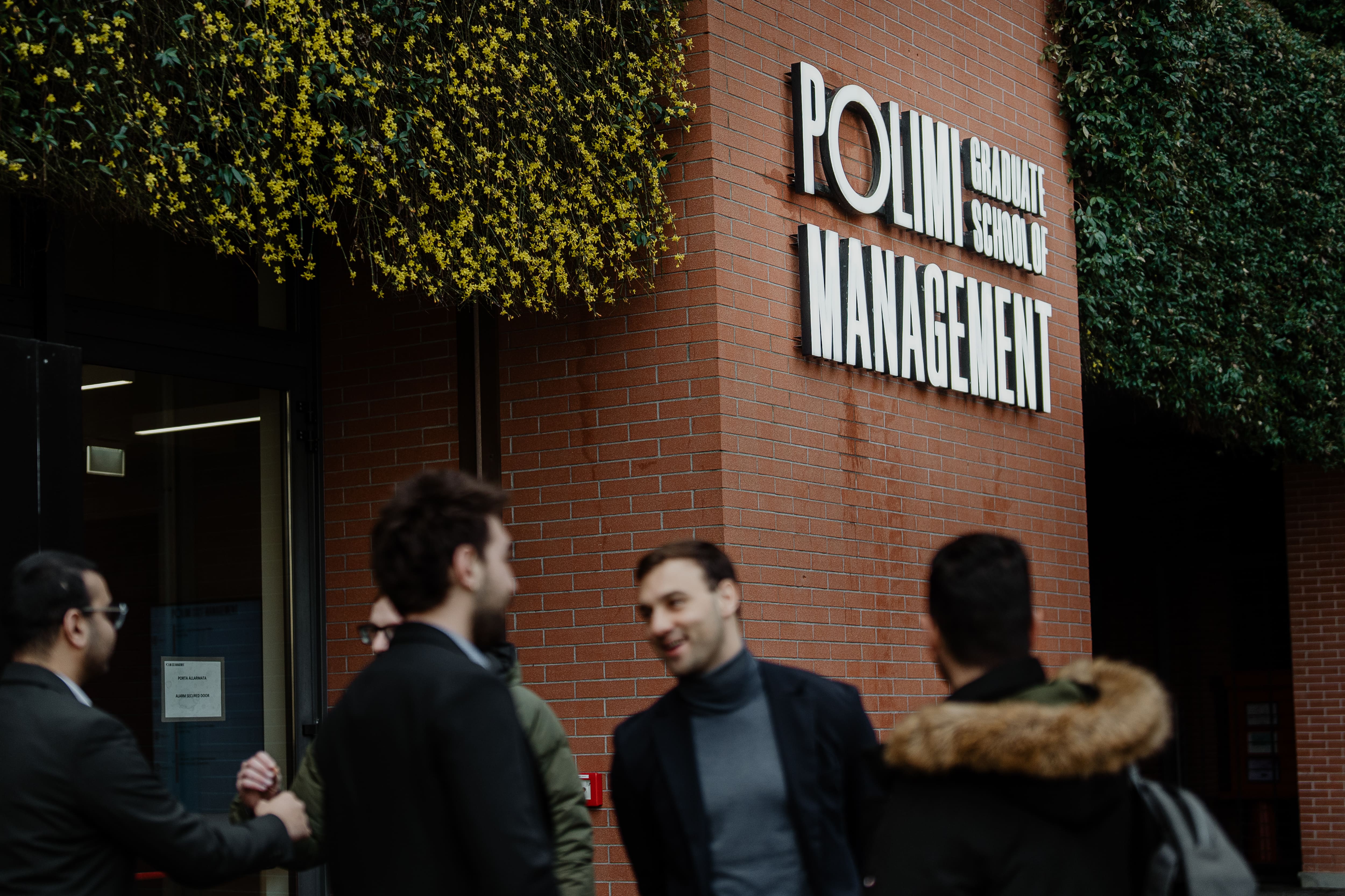 Group of young men greeting each other outside the Polimi Graduate School of Management building with brick walls and greenery.