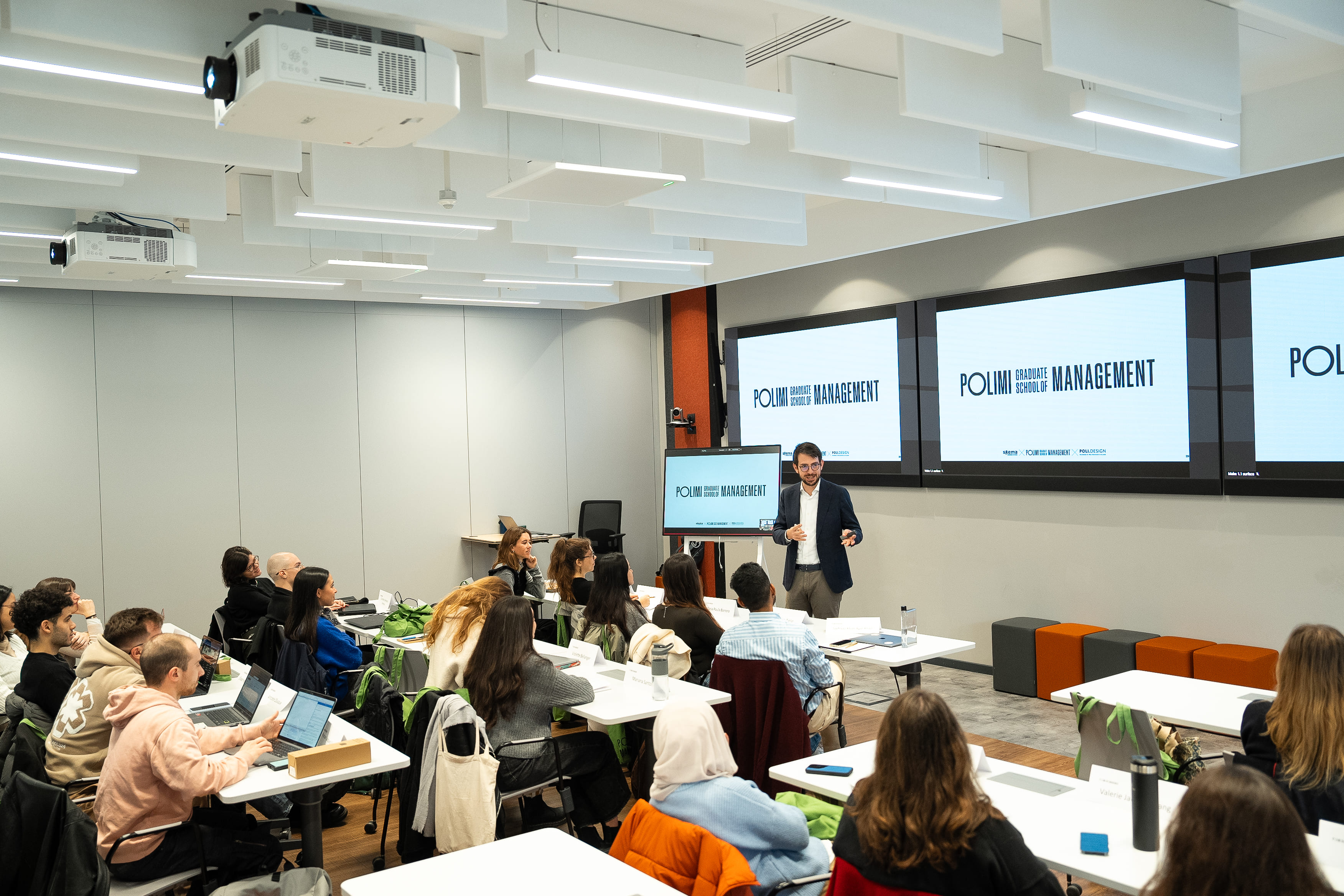 A man speaking to a diverse group of students seated at desks in a modern classroom with multiple screens displaying 'POLIMI Graduate School of Management.'