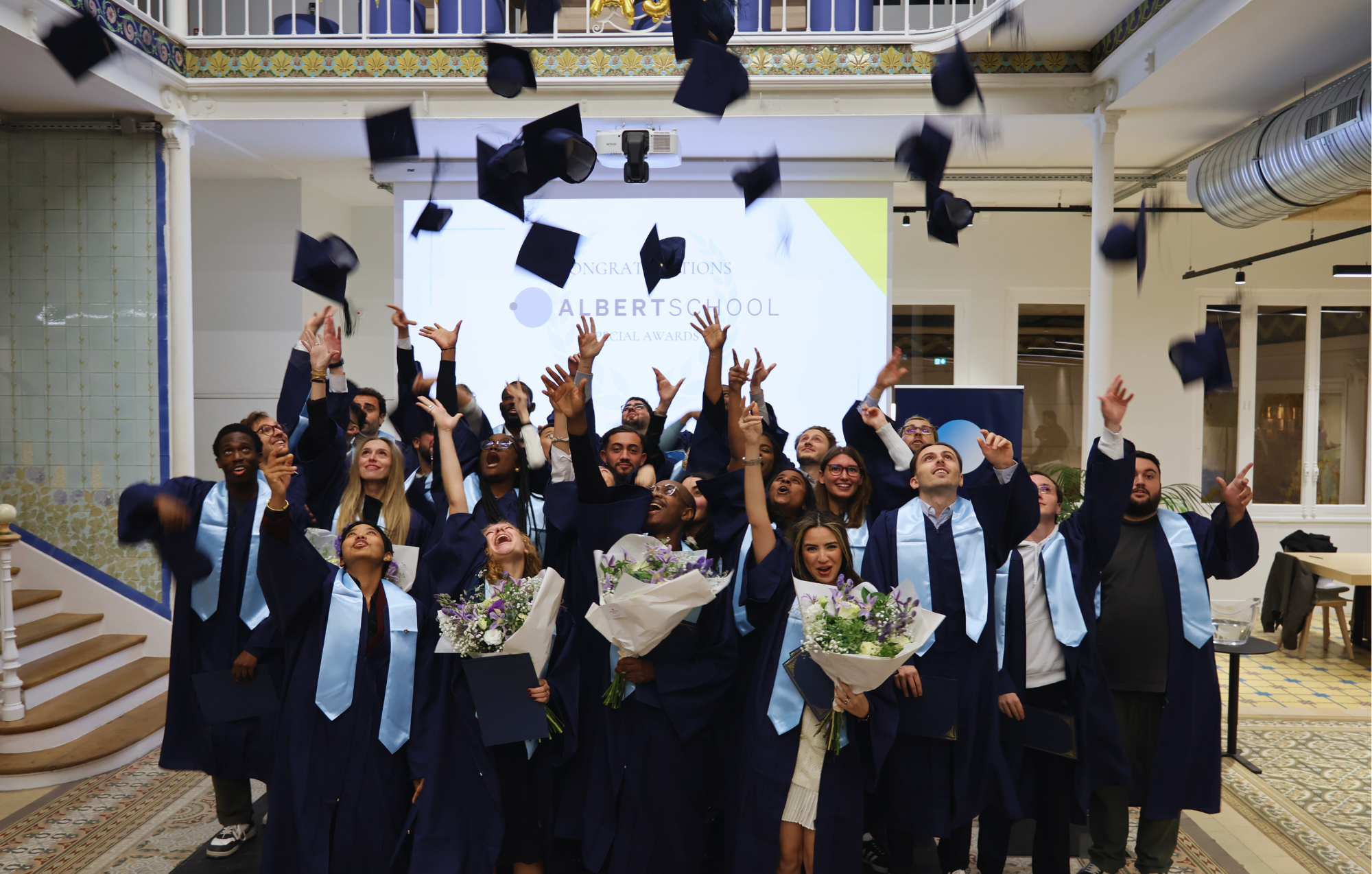 Group of graduates in blue gowns and light blue stoles joyfully throwing their graduation caps in the air indoors with flowers and certificates.