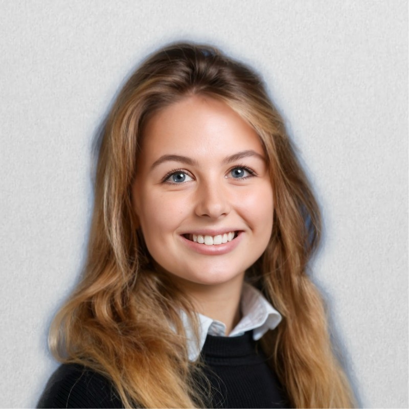 Smiling young woman with long blonde hair wearing a white collared shirt and black sweater against a light gray background.
