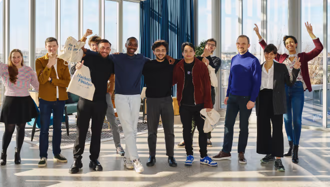 Group of ten diverse young adults standing indoors in front of large windows, smiling and celebrating with some raising their arms.