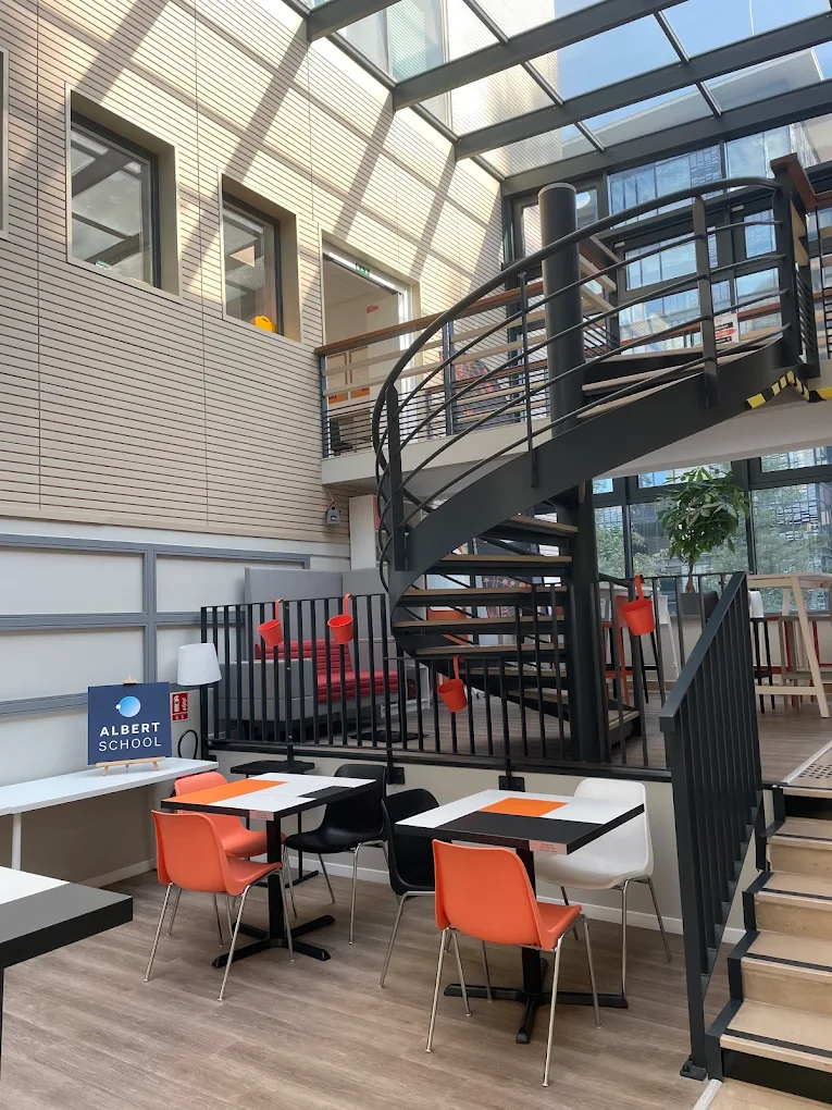 Modern indoor space with spiral staircase, tables with orange, black, and white chairs, and a sign reading Albert School.