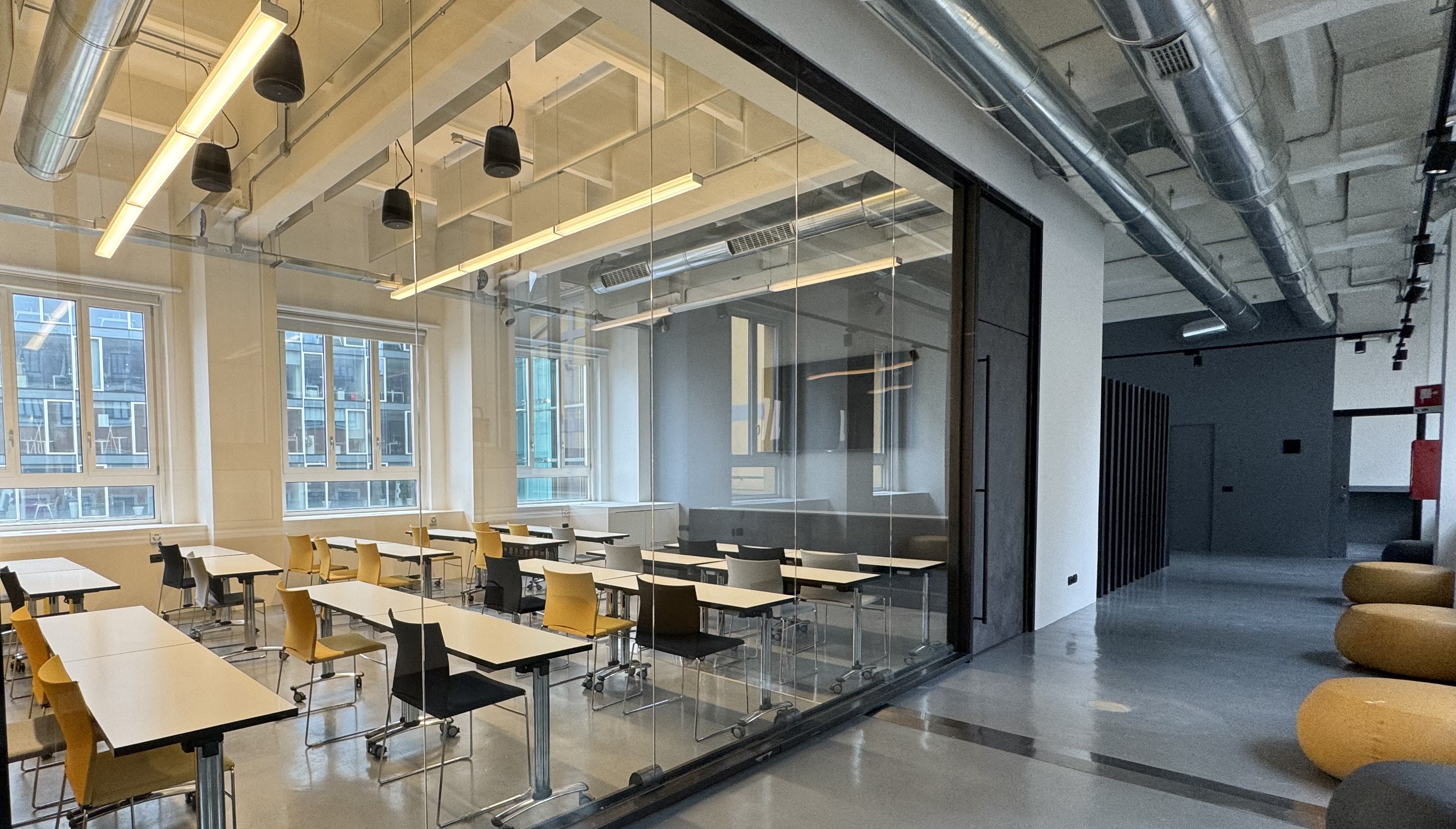 Modern classroom with glass walls, white desks, mixed yellow and black chairs, and a hallway with round seating.