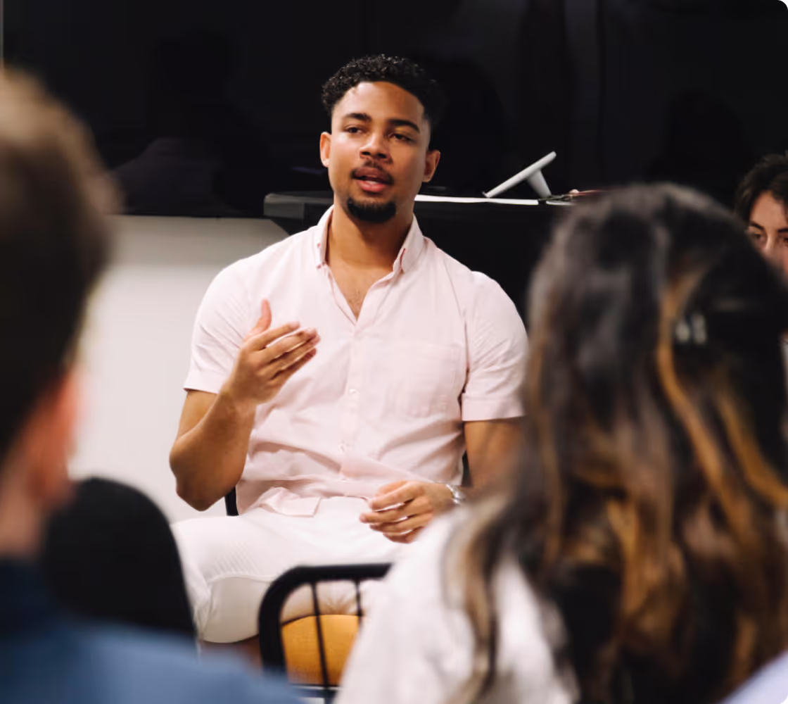 Young man in a light pink shirt speaking to an audience in a discussion setting.