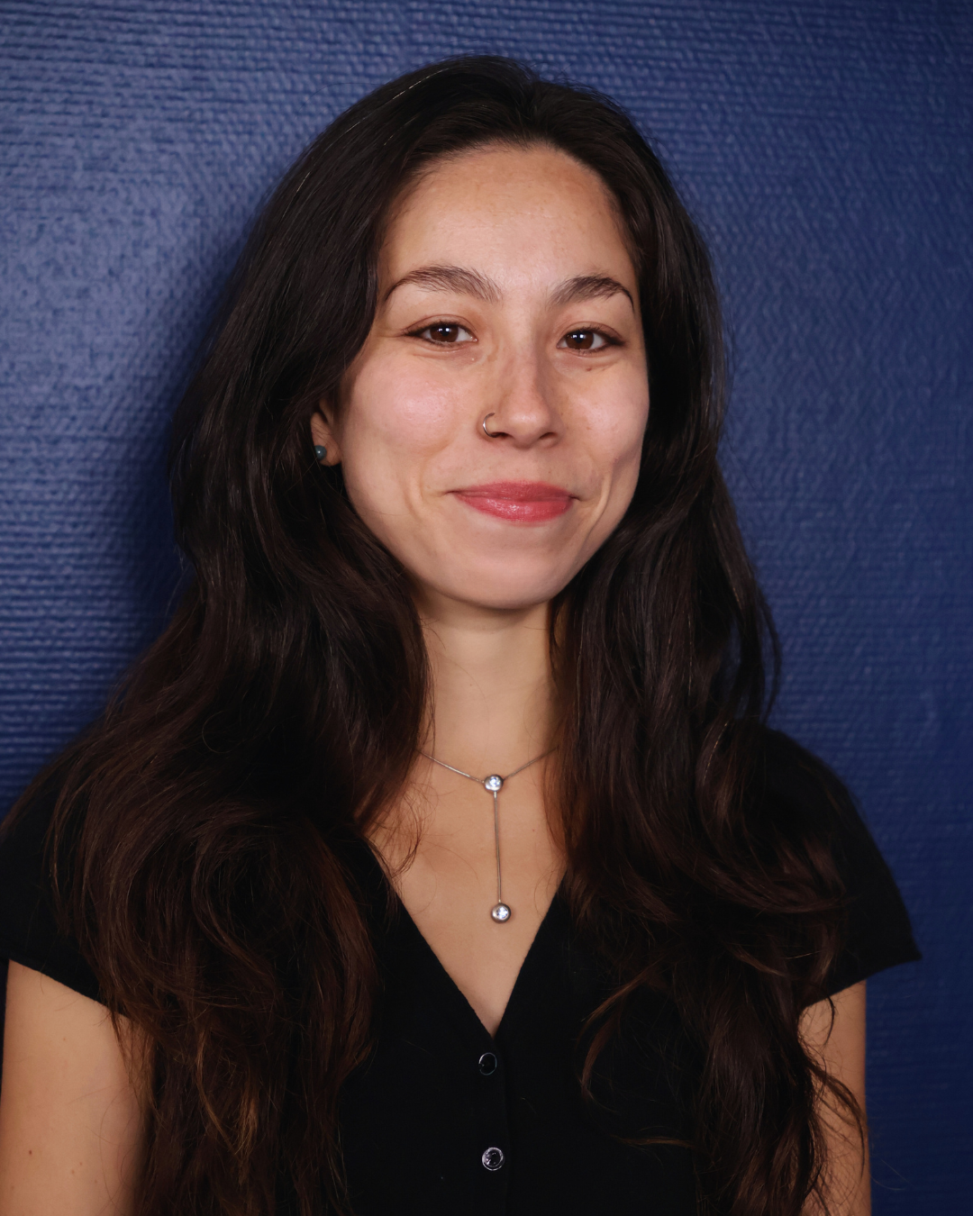 Smiling woman with long dark hair wearing a black top and silver necklace against a blue textured background.