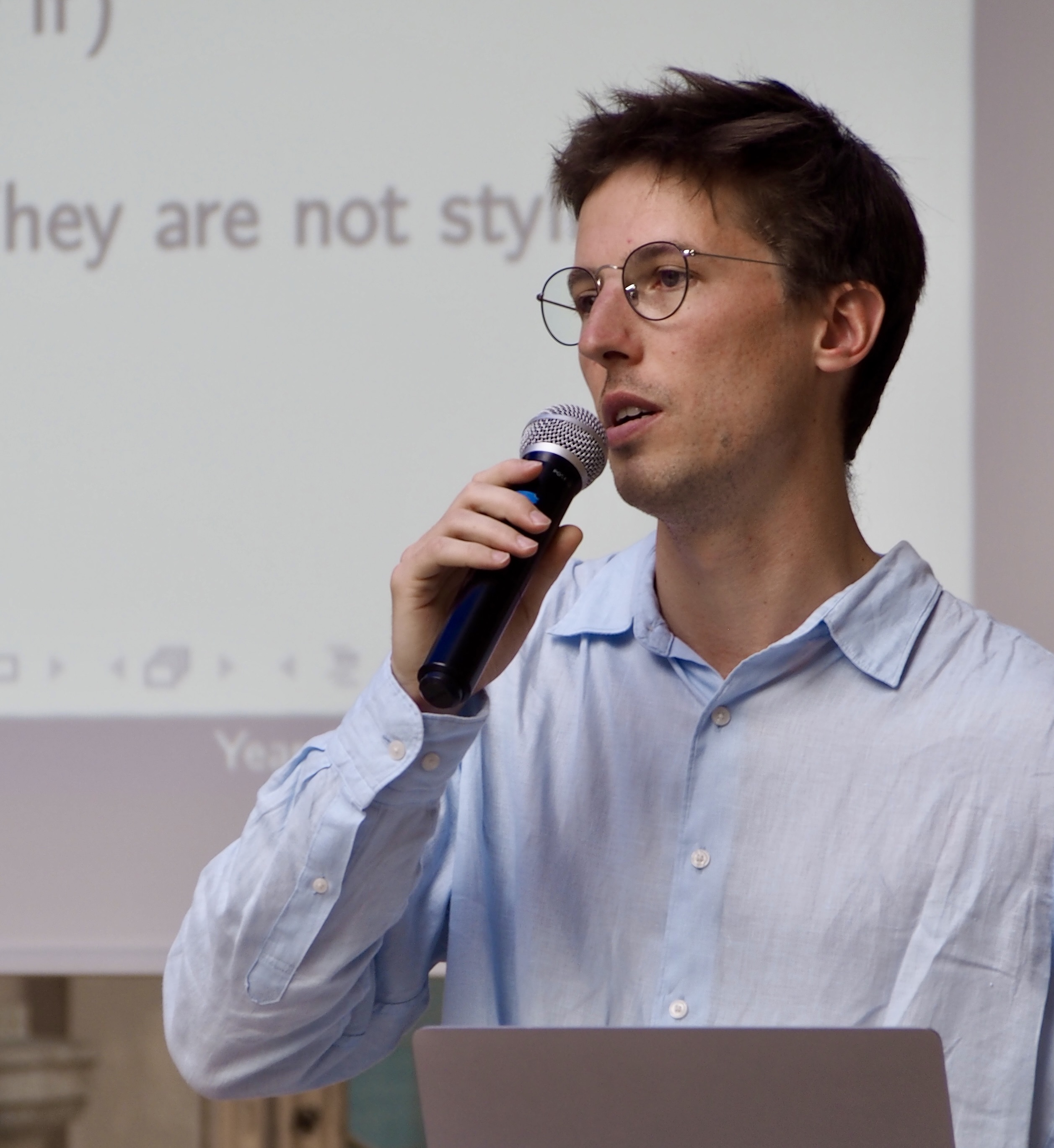 Young man with glasses speaking into a microphone during a presentation, with projection screen in the background.