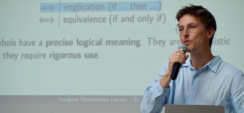Man with glasses speaking into a microphone in front of a projected slide about logical implication and equivalence.