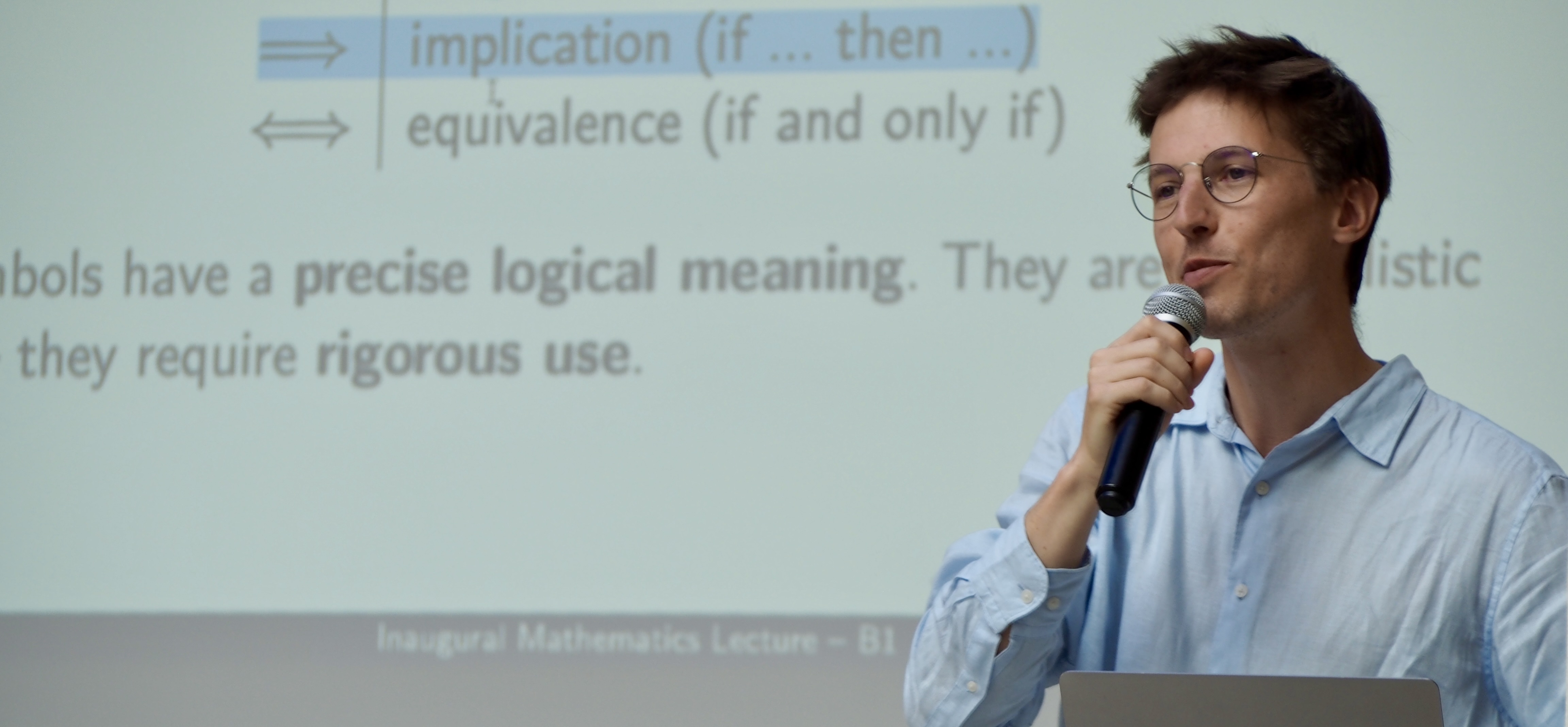 Man with glasses speaking into a microphone in front of a projected slide about logical implication and equivalence.