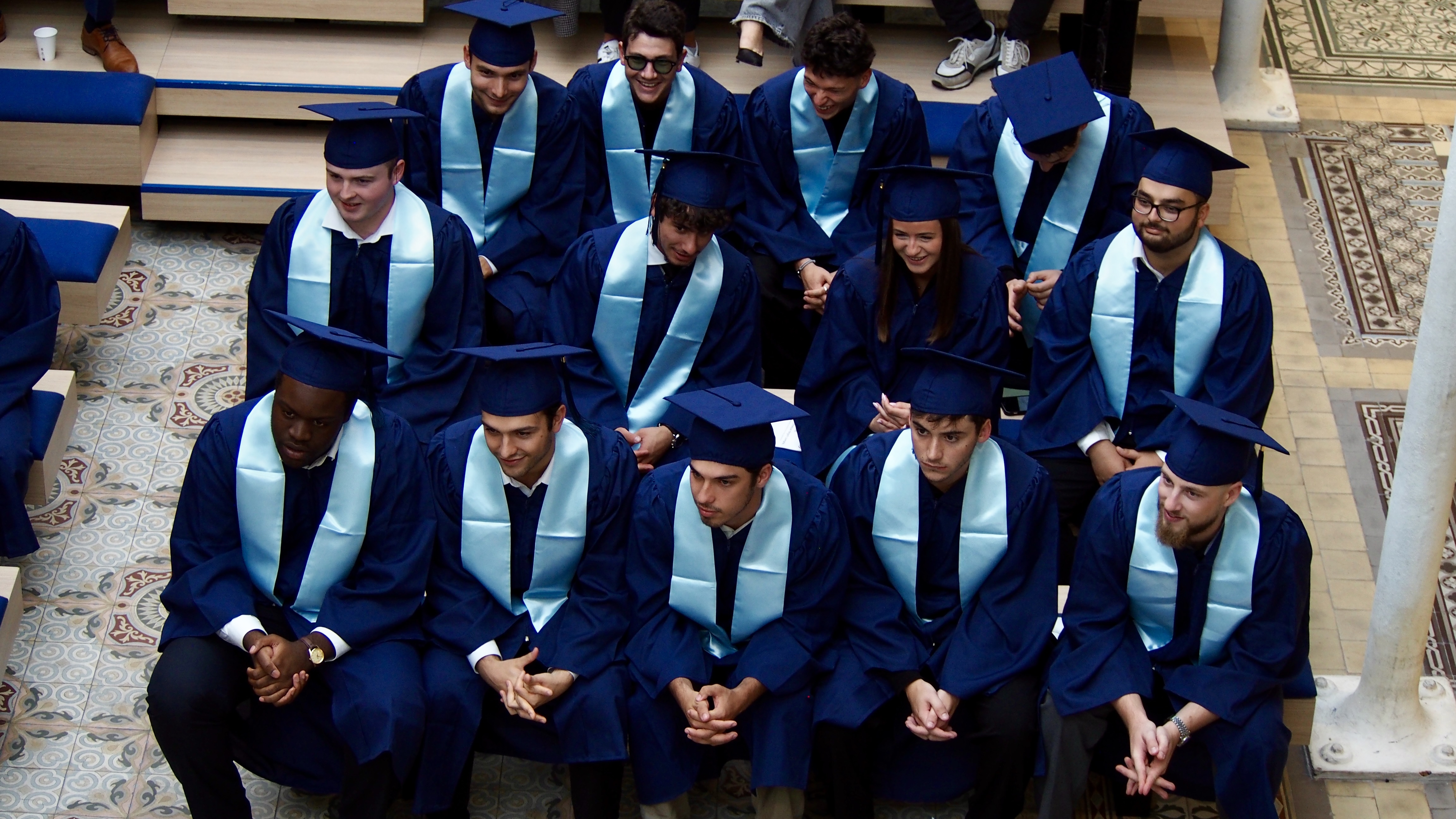 Group of graduates in blue caps and gowns with light blue stoles seated in an indoor venue with patterned tile flooring.