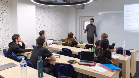 Instructor standing at the front of a classroom giving a lecture to students seated at desks with laptops and notebooks.