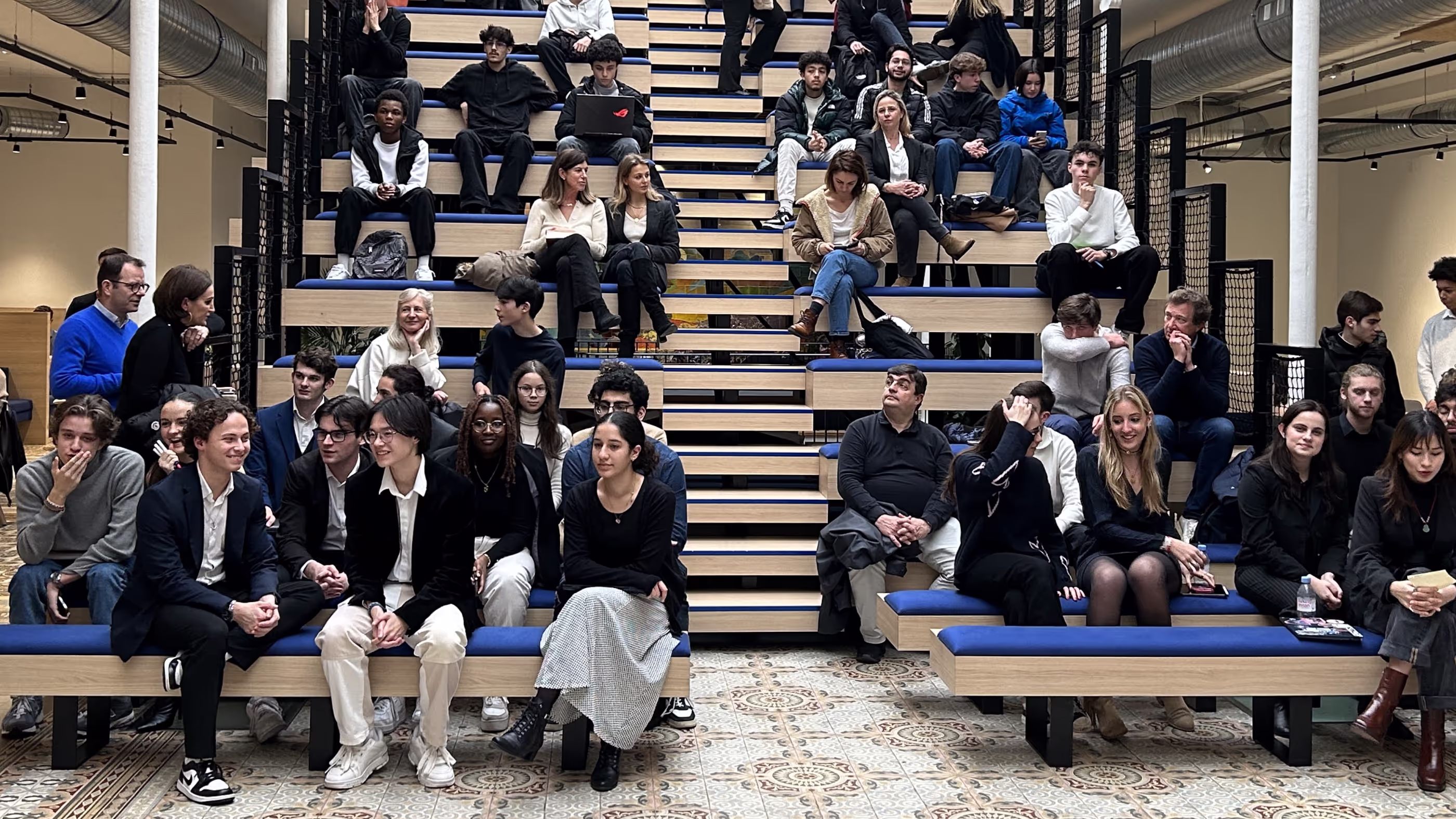 A diverse group of people seated on tiered wooden benches with blue cushions in an indoor space with patterned tile floor and industrial ceiling.
