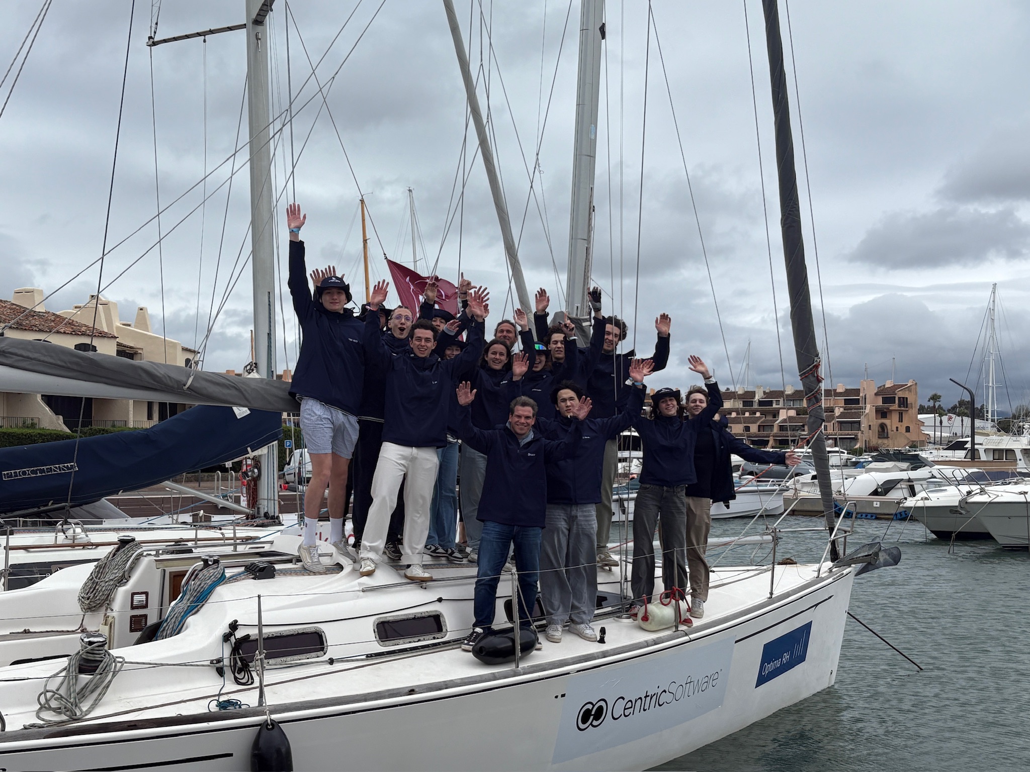 Group of people wearing matching jackets standing and smiling on a sailboat docked at a marina.