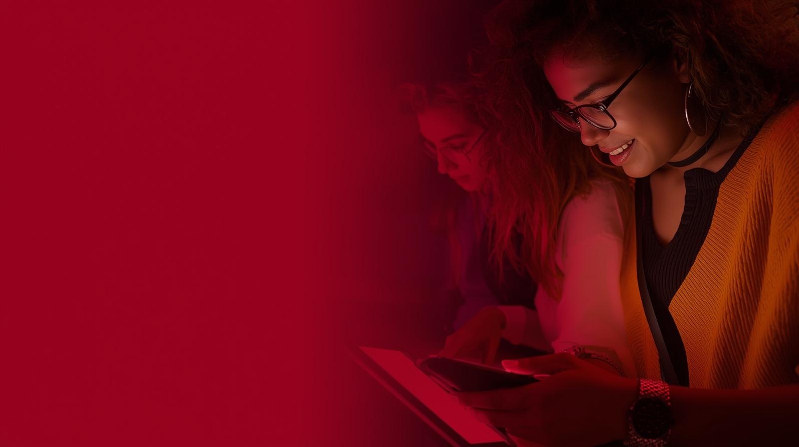 Two women sitting closely, one smiling while using a smartphone and the other focused on a tablet, both illuminated by red light.