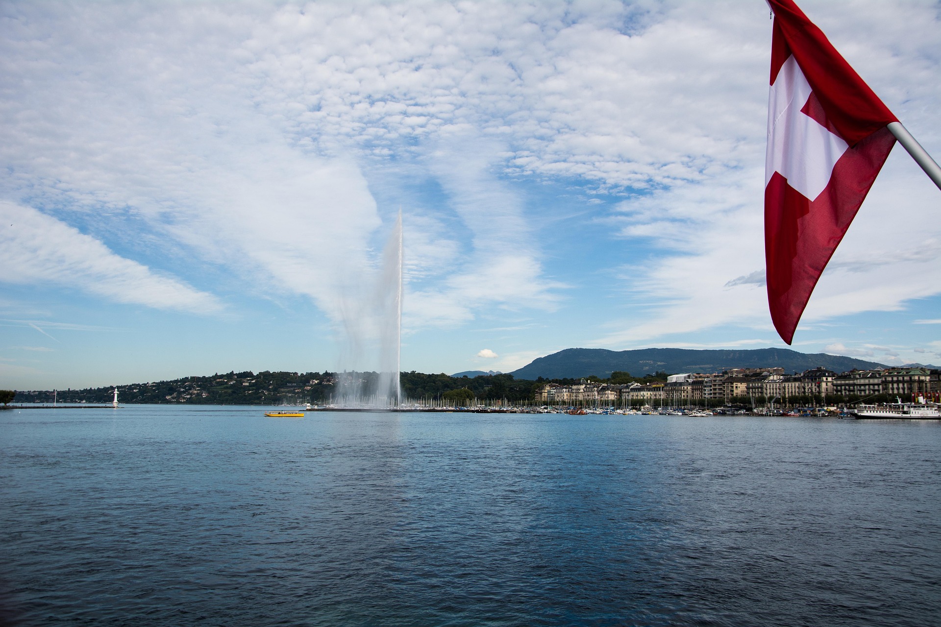 Lake with a large water jet fountain, buildings and mountains in the background, and a Swiss flag on the right.