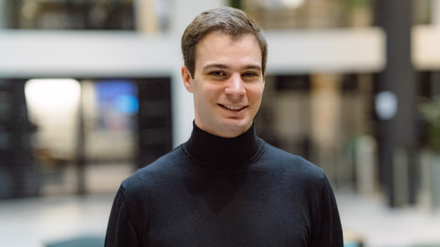 Smiling young man wearing a black turtleneck sweater in a blurred indoor setting.