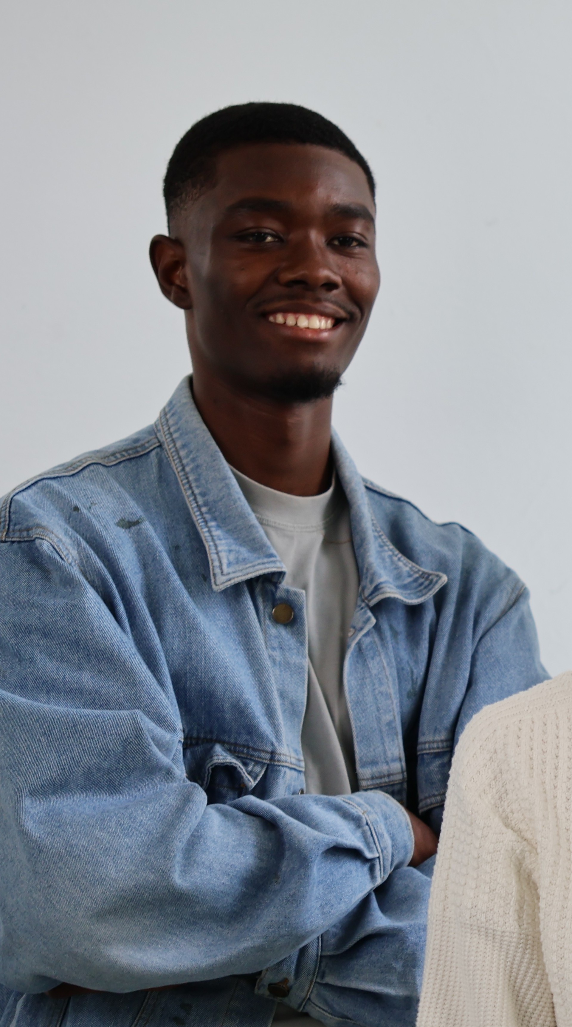 Smiling young man with a short haircut wearing a blue denim jacket over a light shirt against a plain light background.