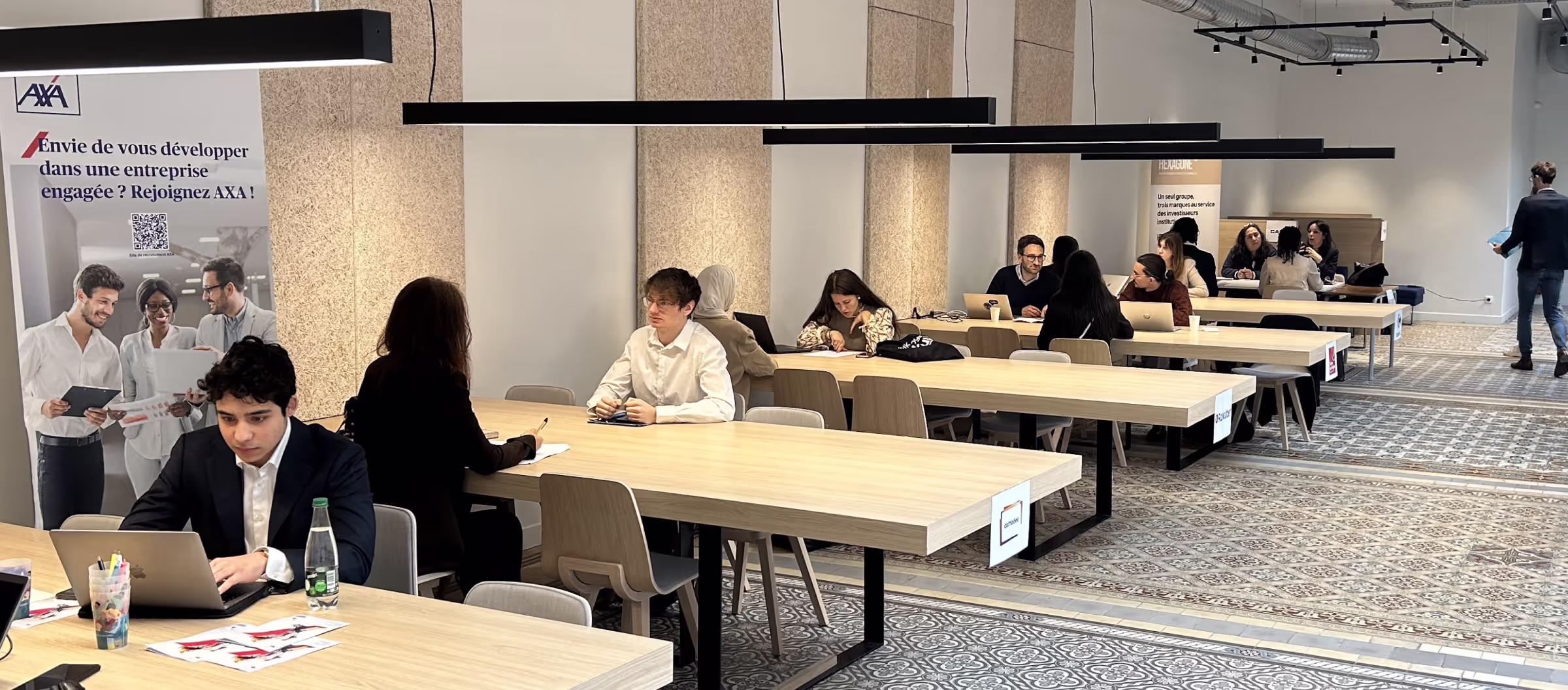 Modern open workspace with multiple light wooden tables where people are working and discussing in a bright room with patterned tile floor.