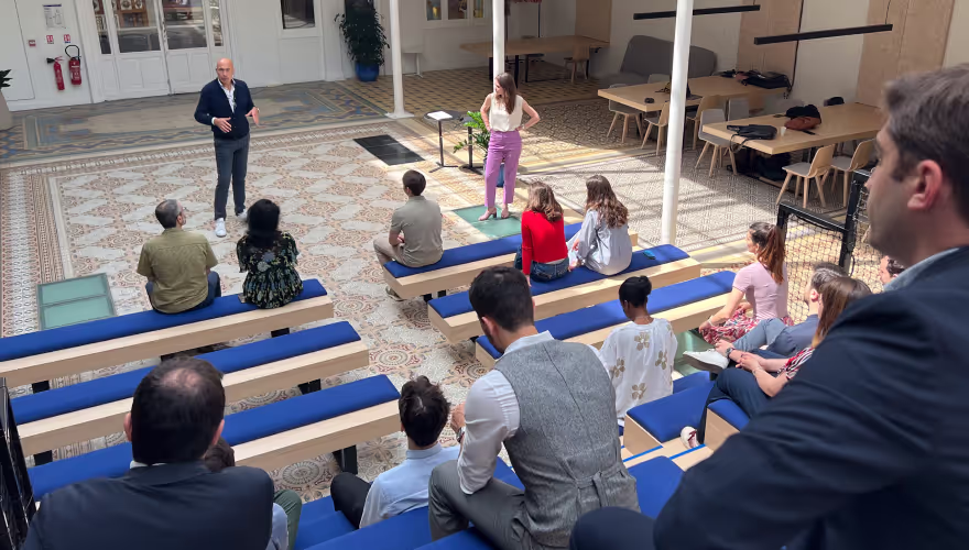 Man in blue jacket speaking to an attentive audience seated on tiered benches in a spacious room with patterned tile floor.