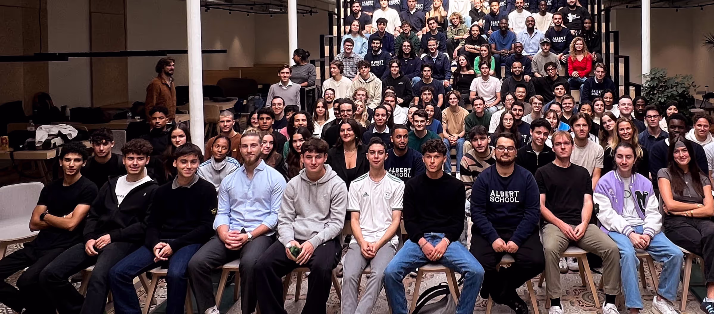 Large diverse group of young adults and students posing for a group photo indoors on tiered seating and chairs.