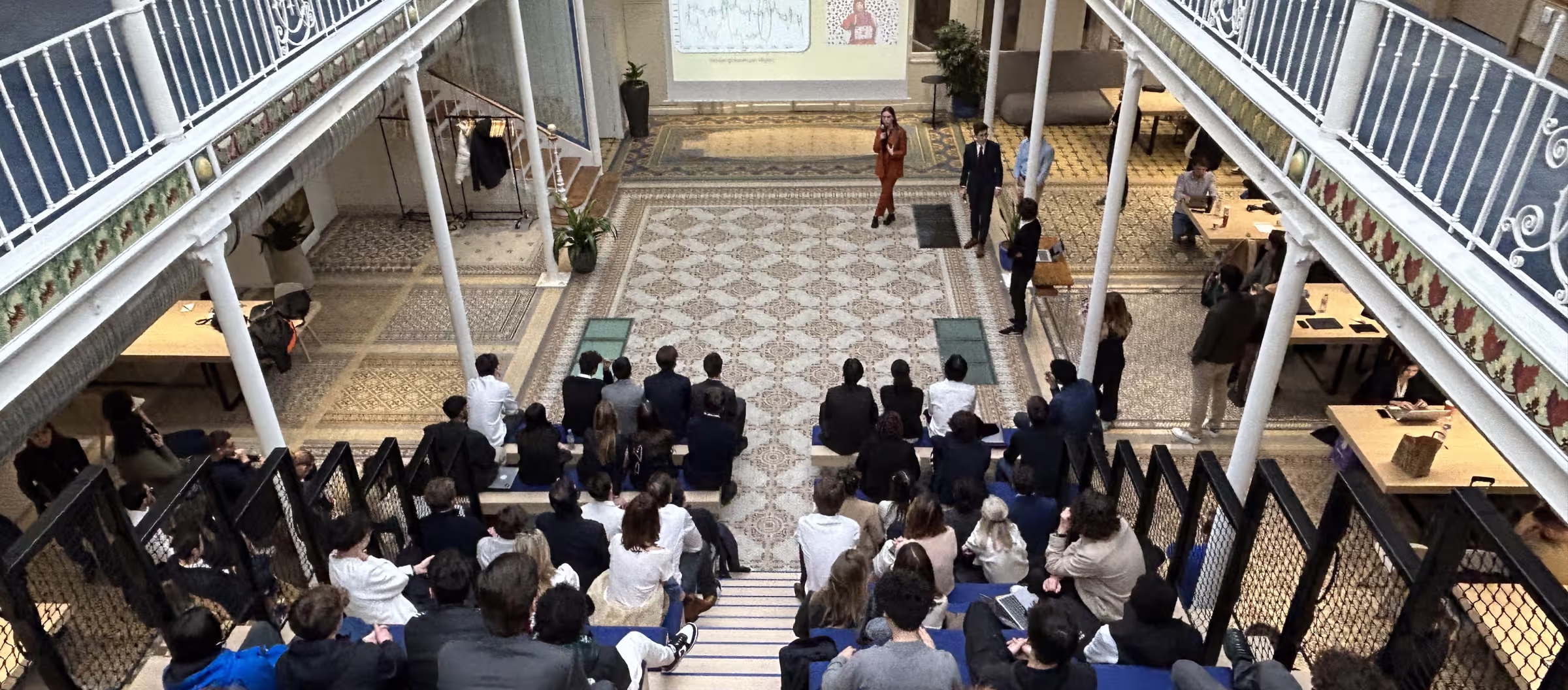 Overhead view of a group of people seated on tiered benches in a courtyard listening to two presenters near a projection screen.