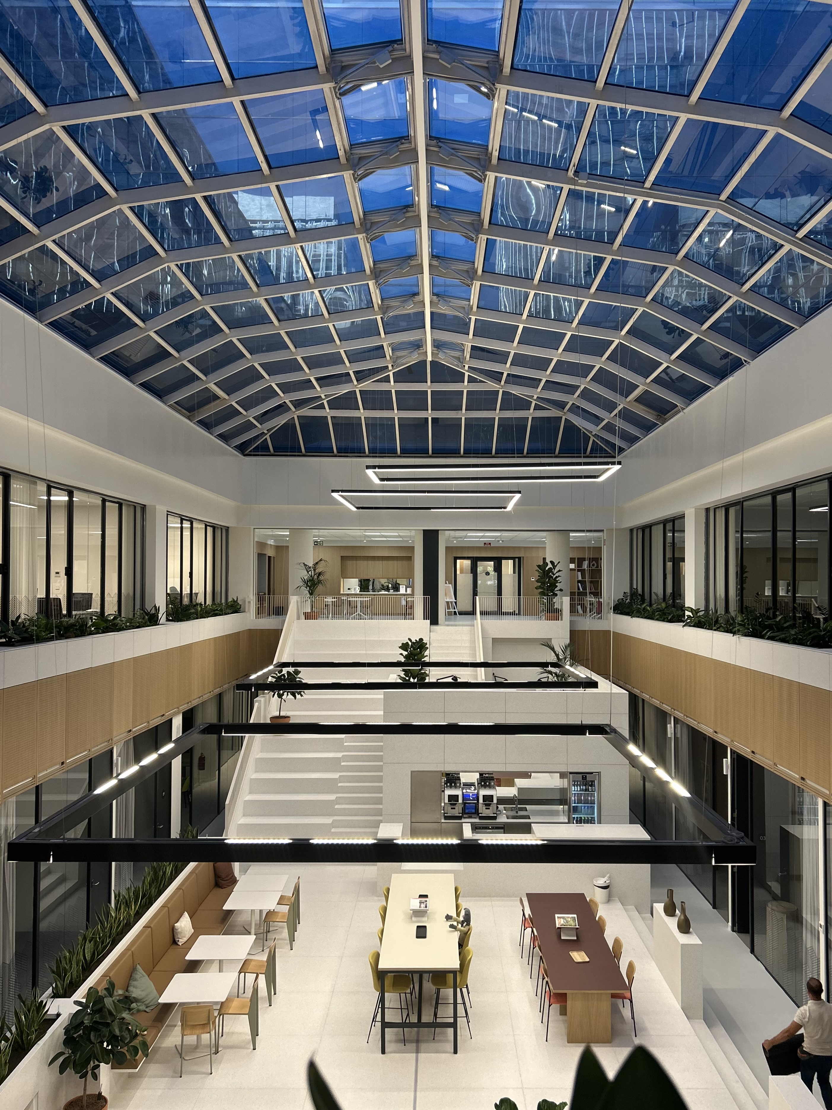 Modern office atrium with glass ceiling, seating areas, tables, plants, and hanging rectangular lights.