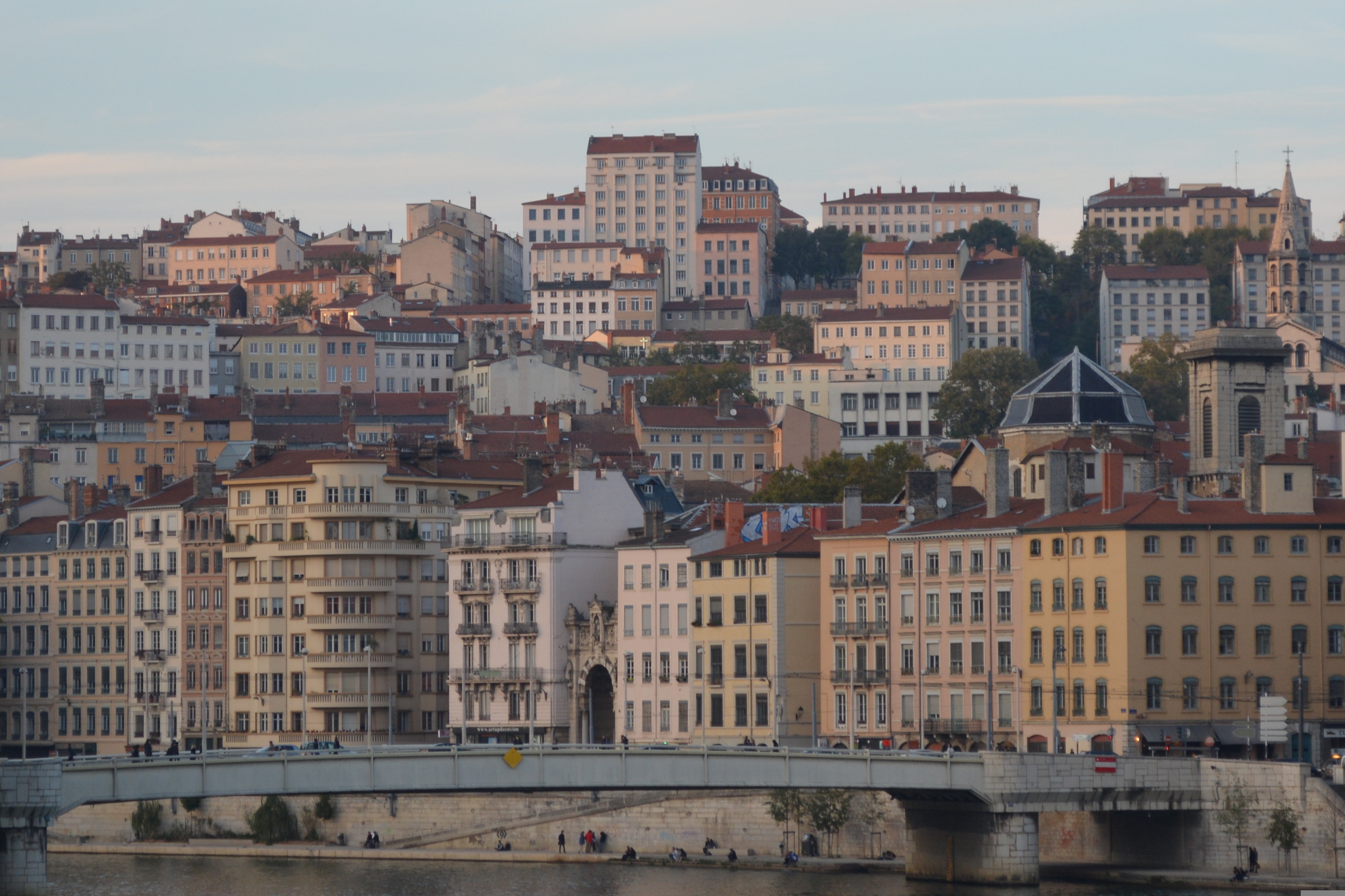 Riverfront view of historic multi-story buildings and residential apartments in Lyon, France, with a stone bridge crossing the river.