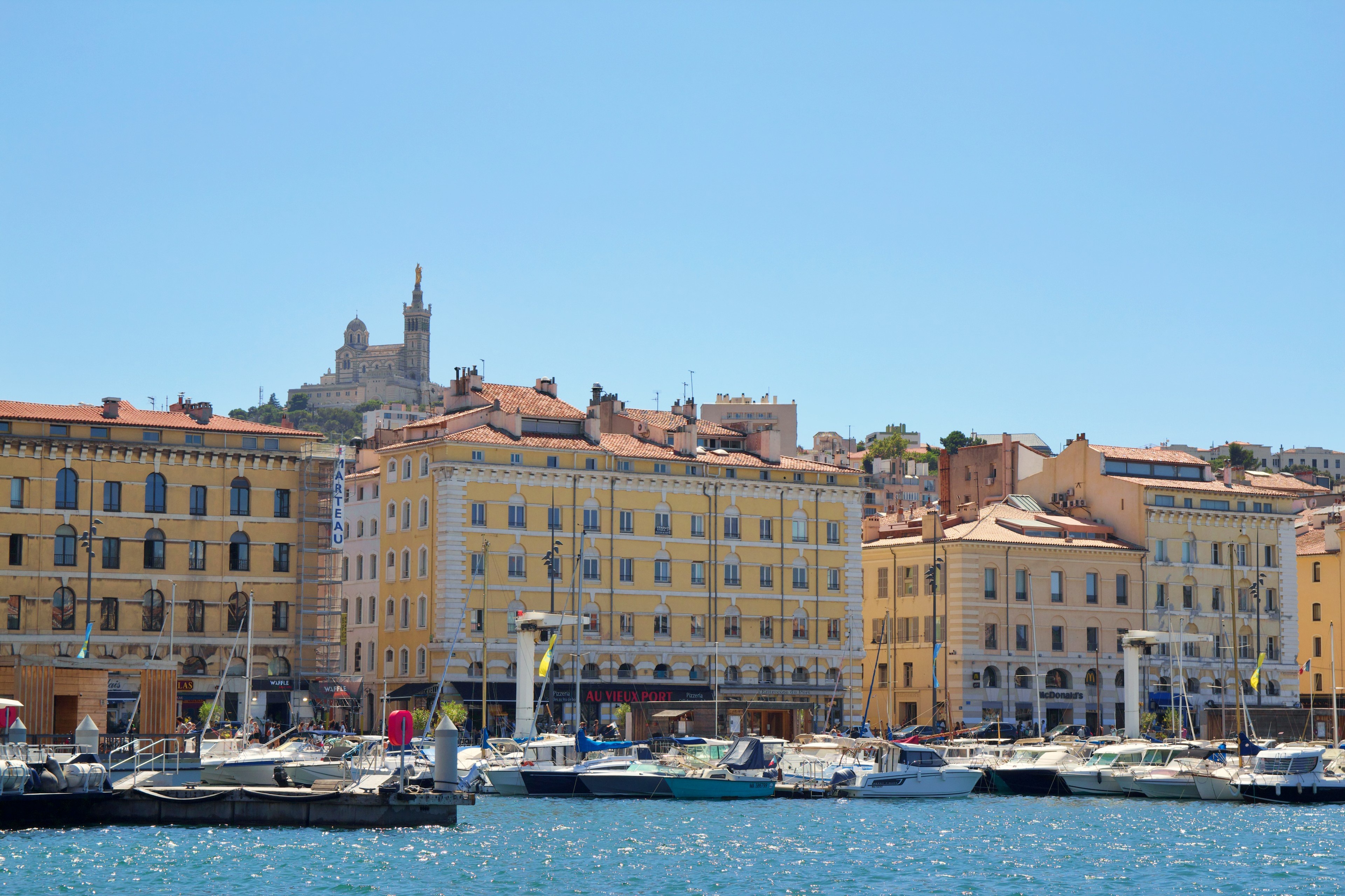 Boats docked at a marina with yellow buildings and the Basilica Notre-Dame de la Garde on a hill in the background under a clear blue sky.