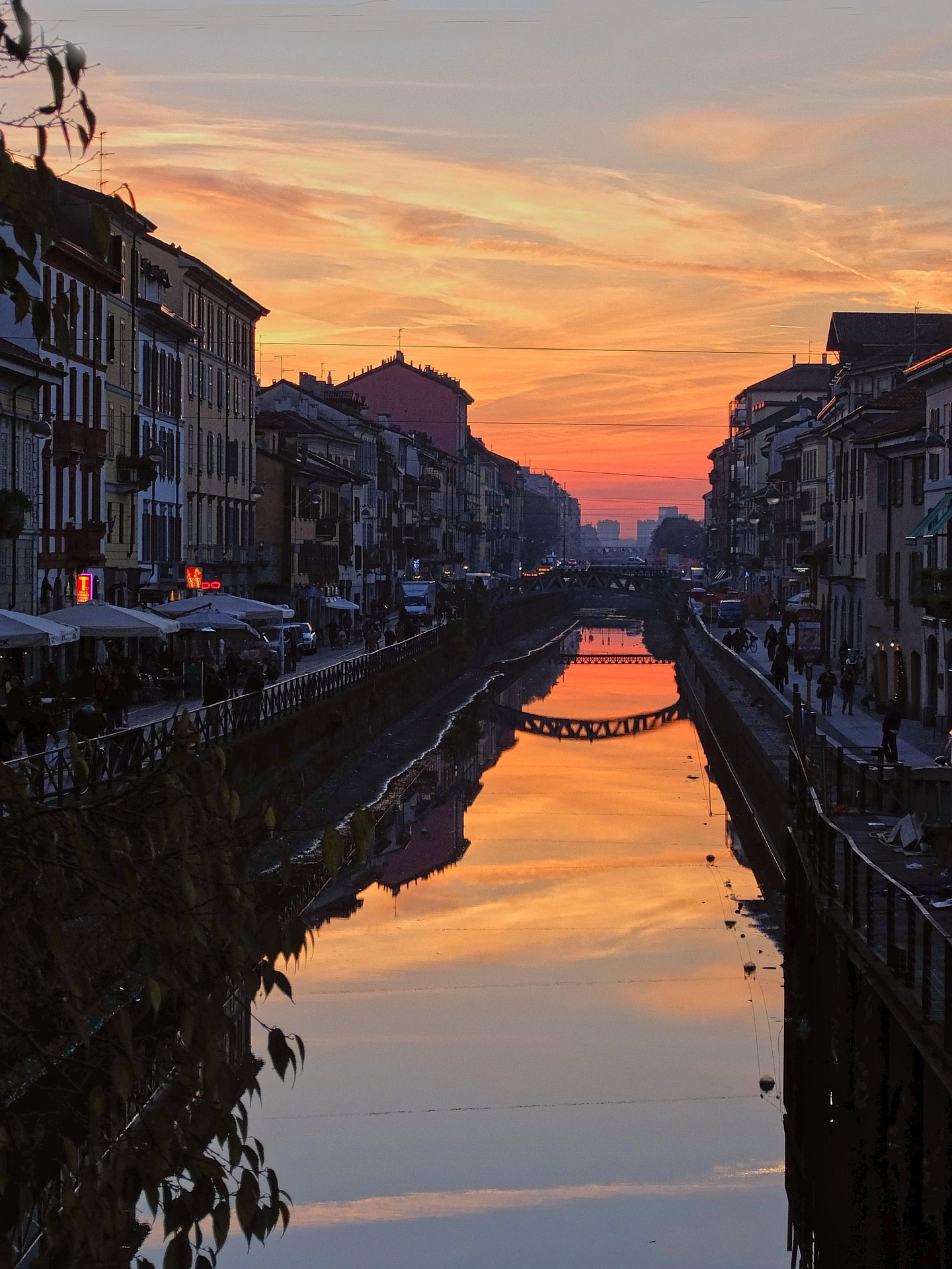 Canal in Milan lined with buildings and pedestrian walkways at sunset reflecting orange and pink colors.
