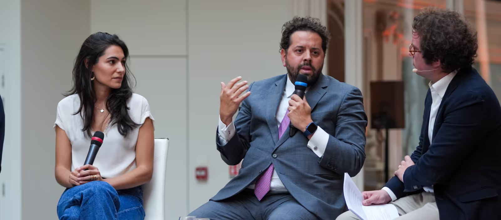 Three people engaged in a panel discussion, with the man in the center speaking into a microphone while the others listen.
