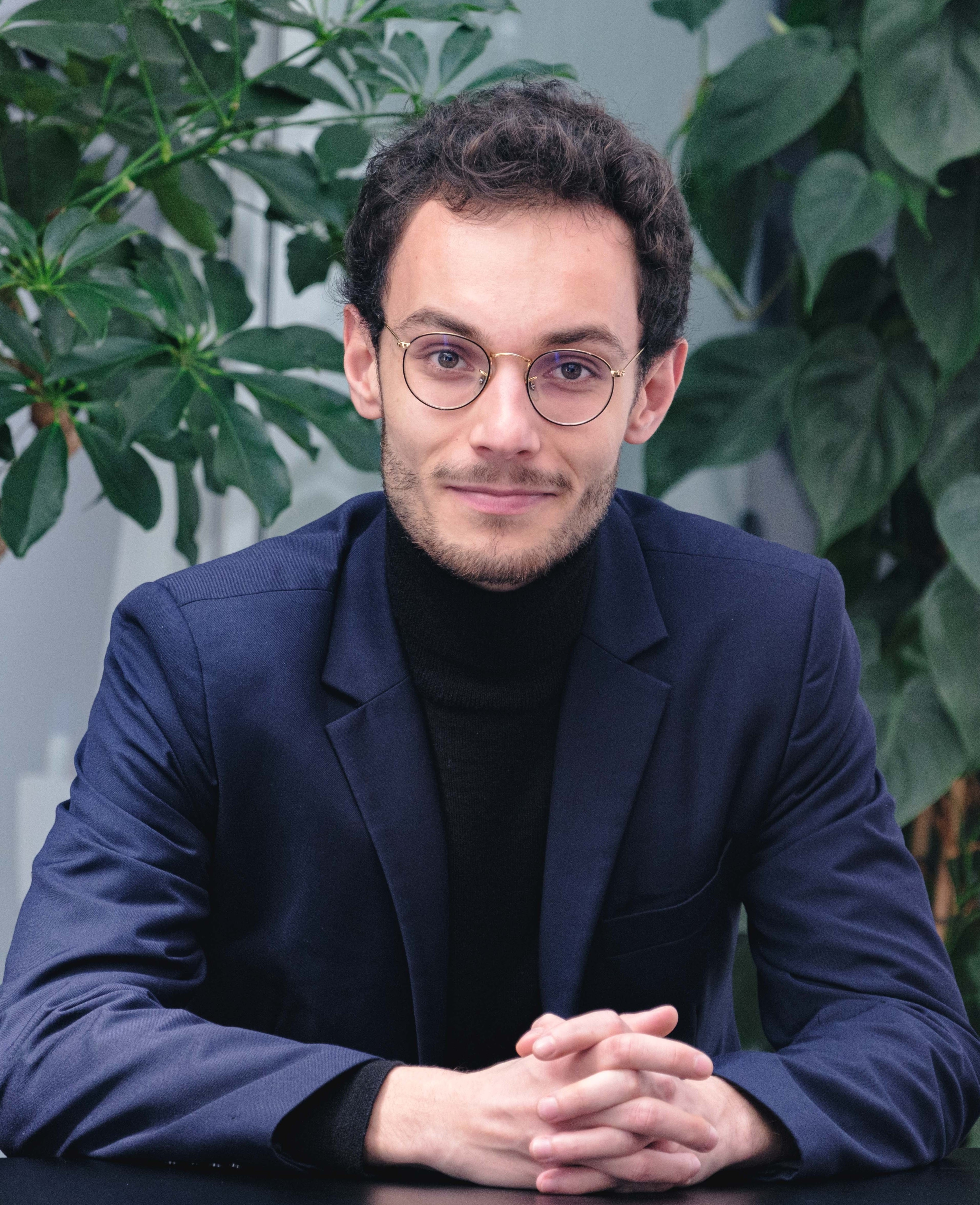 Young man with glasses wearing a dark blazer and black turtleneck, sitting with hands clasped in front of lush green plants.