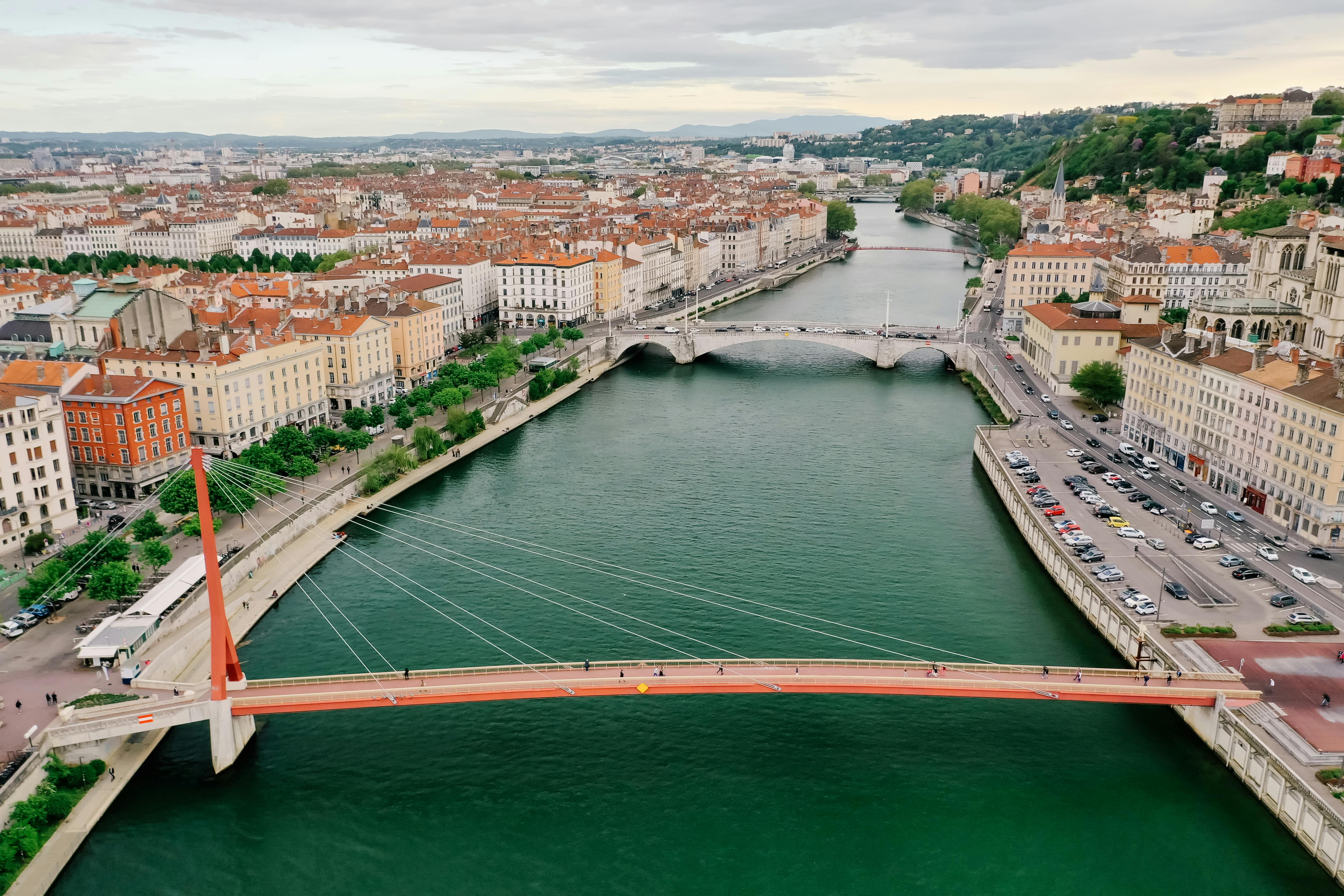 Aerial view of a city river with two bridges, one modern suspension footbridge and one traditional stone bridge, lined with buildings and streets.