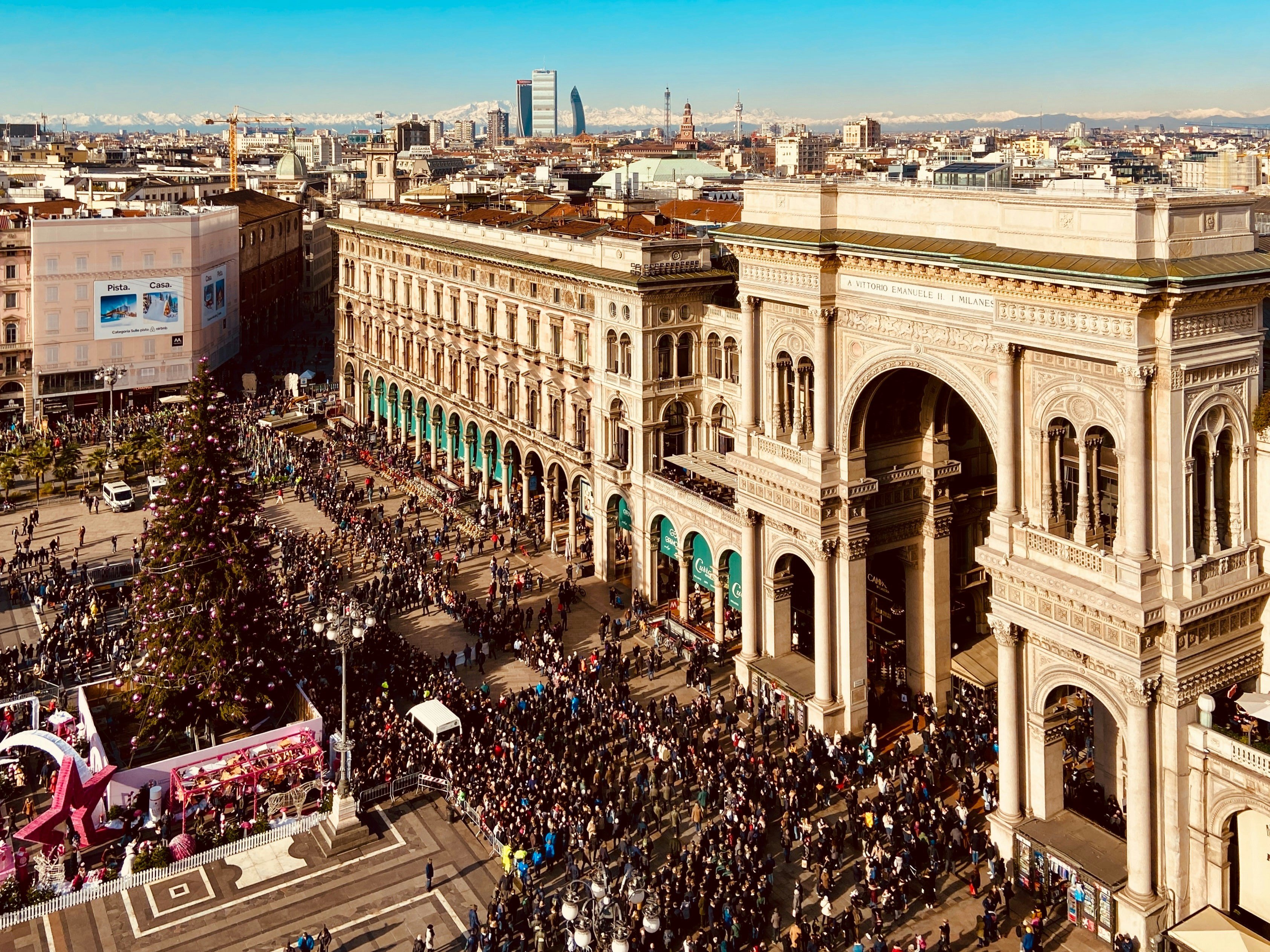 Crowded Piazza del Duomo in Milan with the Galleria Vittorio Emanuele II and a large decorated Christmas tree.