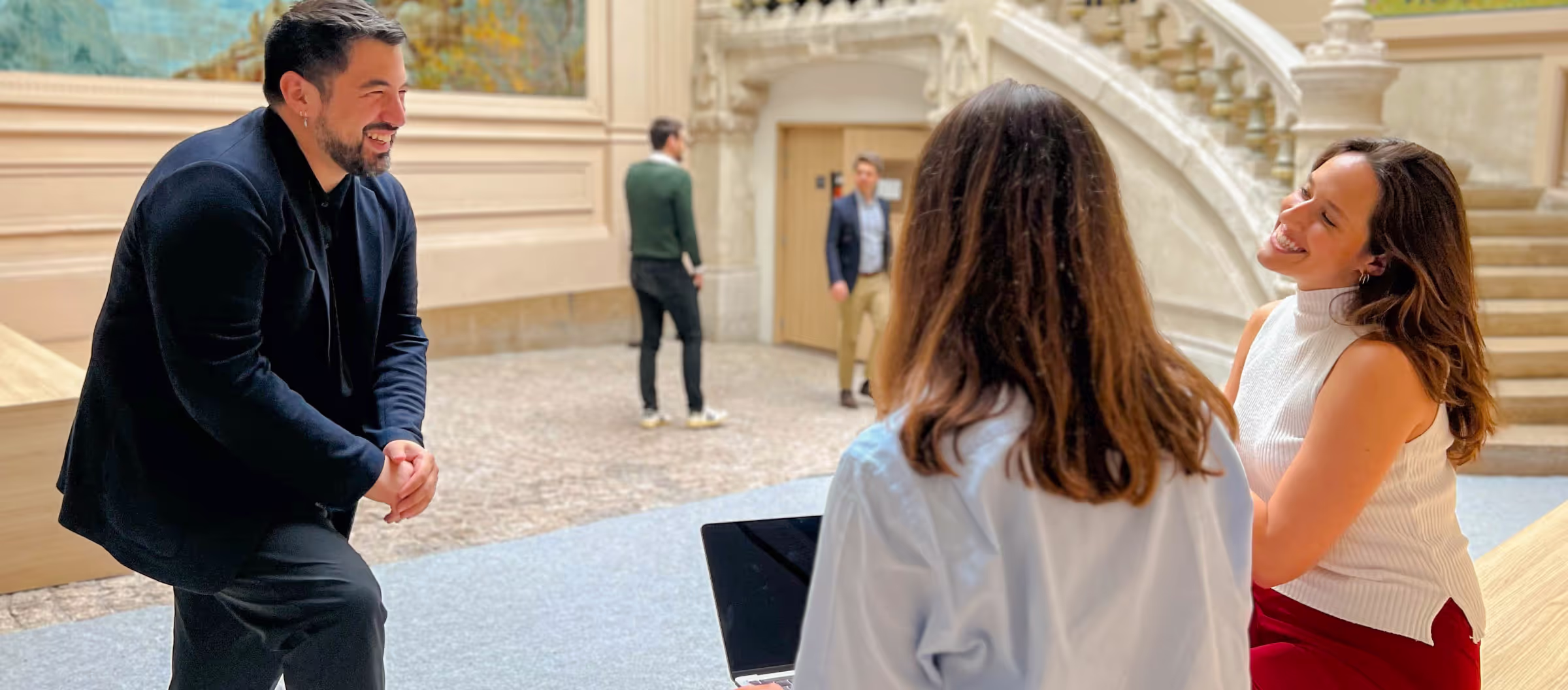 Three people engaged in a lively conversation in a spacious indoor area with stairs and ornate walls.