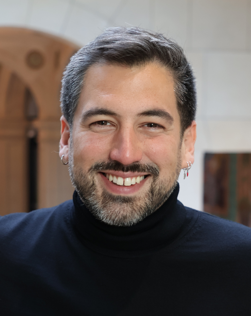 Smiling man with salt-and-pepper hair and beard wearing a black turtleneck and multiple earrings.