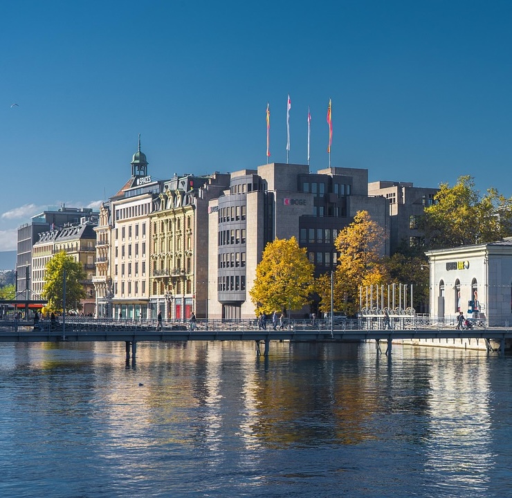 City buildings along a river with a pedestrian bridge and trees with autumn foliage under a clear blue sky.